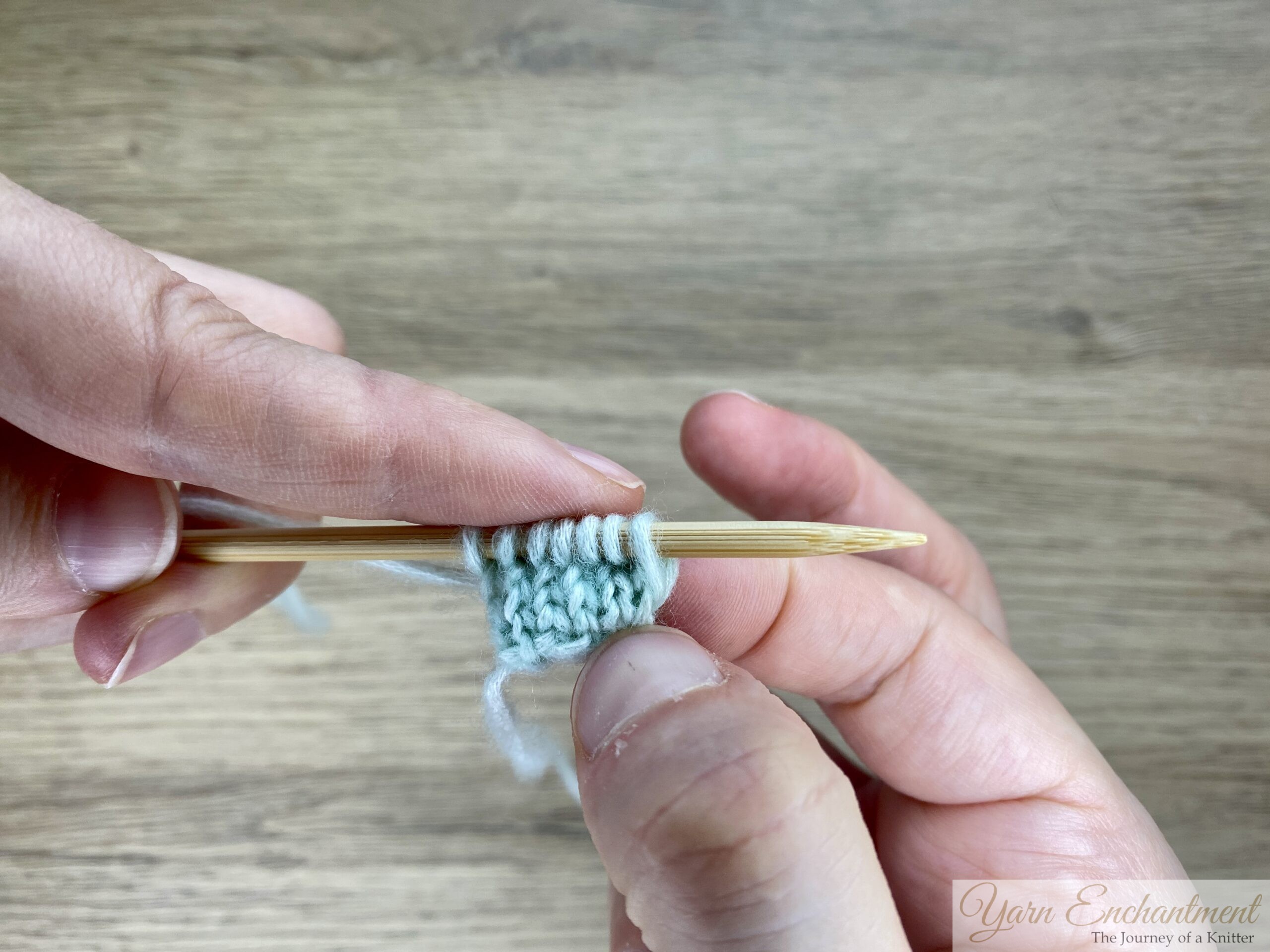 A close-up of a hand holding a bamboo knitting needle with mint green stitches, displaying the rounded and neat edge of the knitted I-cord.