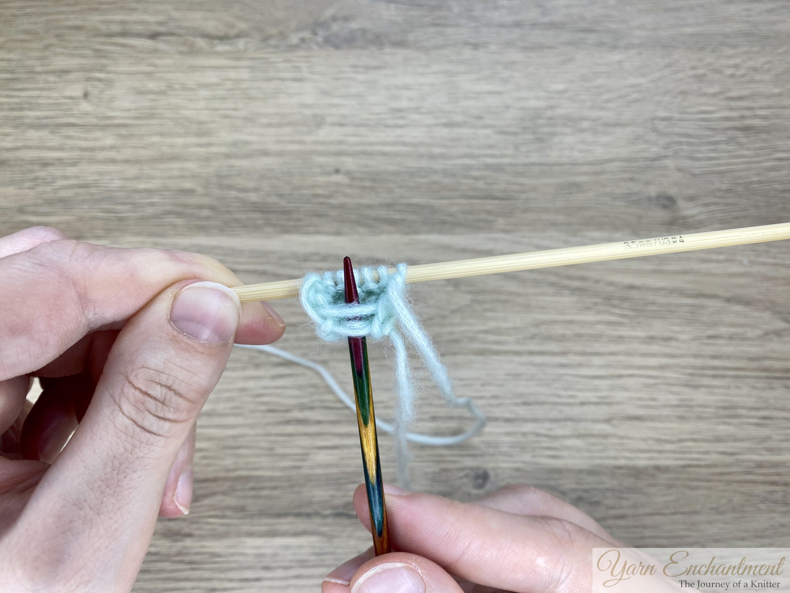 Hands holding two knitting needles with a row of mint green stitches, showing the formation of a wide I-cord segment worked over seven stitches.