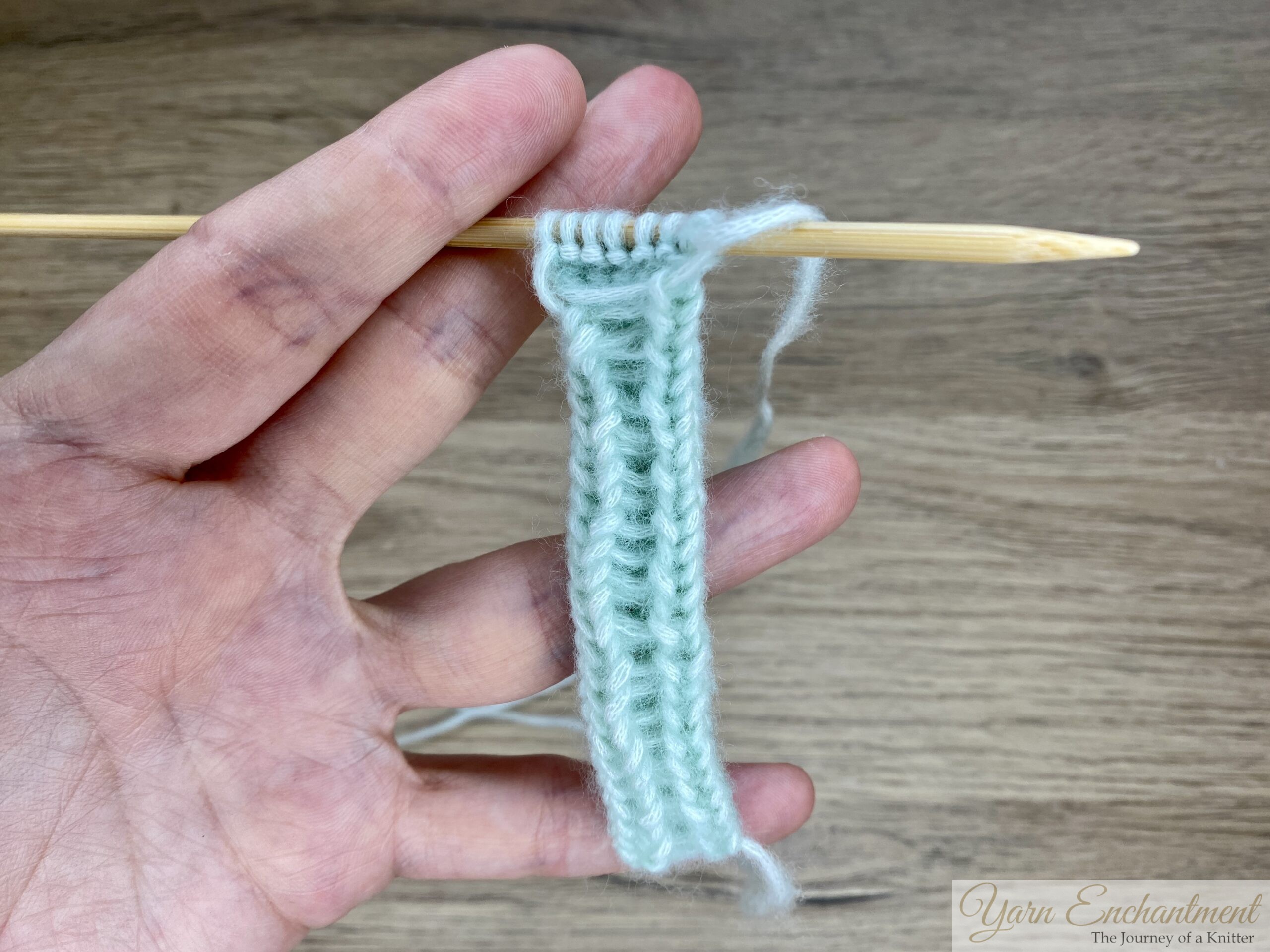 The same I-cord, held on a knitting needle by both hands. The stitches are arranged in a row on the needle, clearly demonstrating the open tube formation.