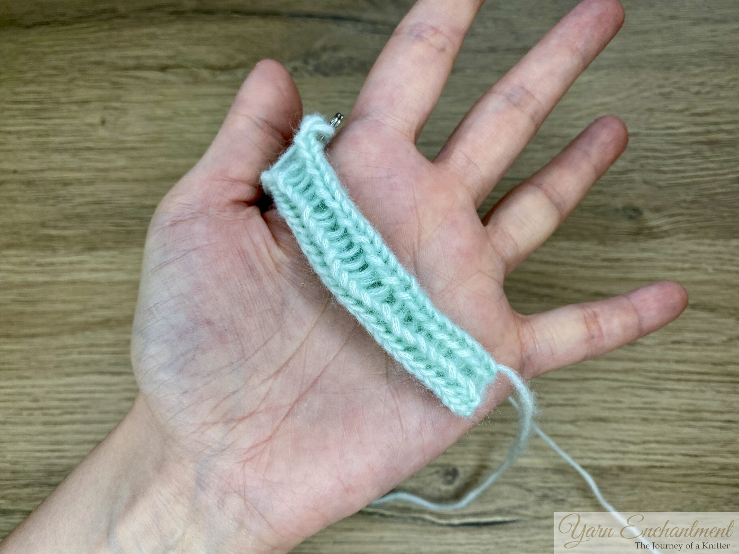 A 7-stitch I-cord in mint green yarn is displayed across an open hand. The knitted tube is clearly visible, showing its width and structure before being closed.