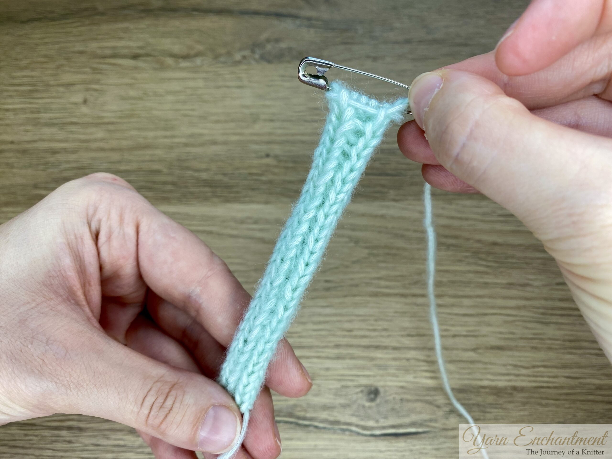 The I-cord is held by both hands, with one end secured using a safety pin. Showing the I cords back after rolling it in the hand