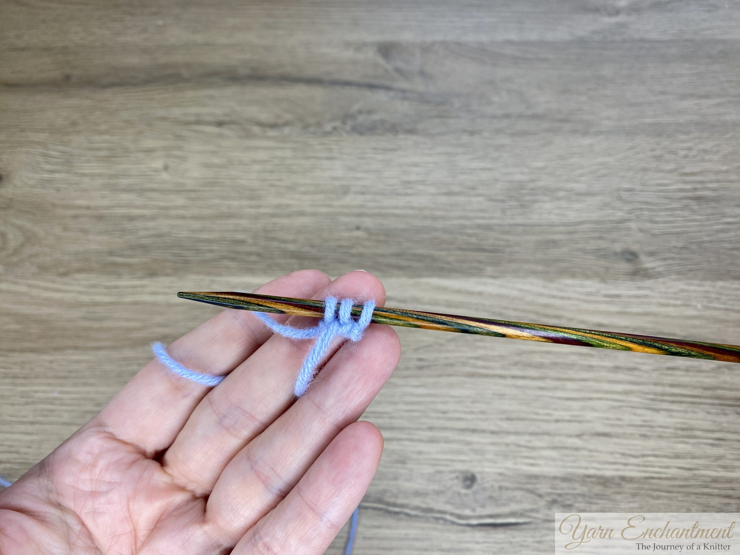 Hand holding three stitches of light blue yarn cast onto a wooden knitting needle, ready to begin an i-cord.