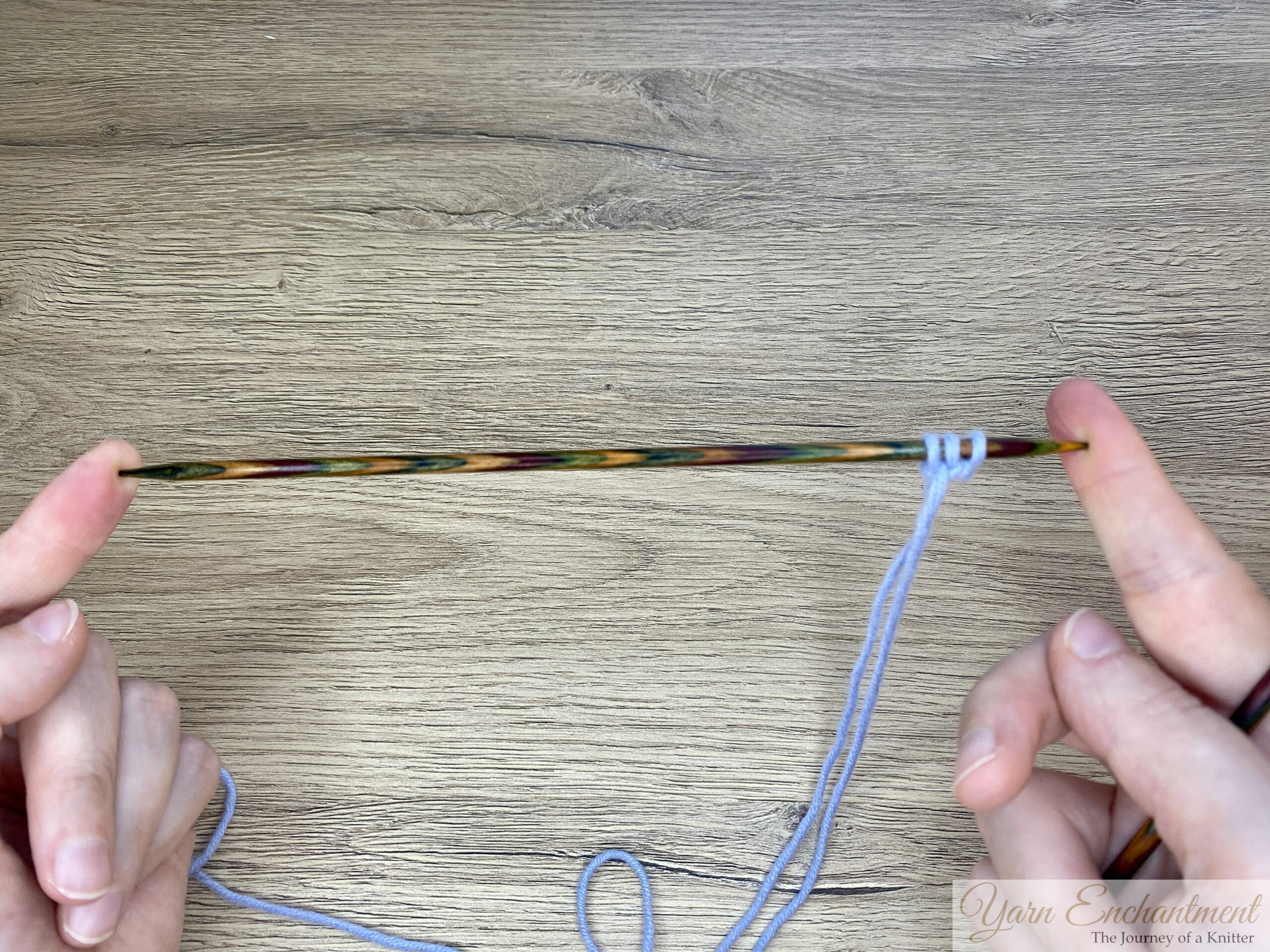 Both hands holding a wooden knitting needle with three stitches of light blue yarn, demonstrating how to slide stitches to the end of the needle before knitting the next row in i-cord technique.