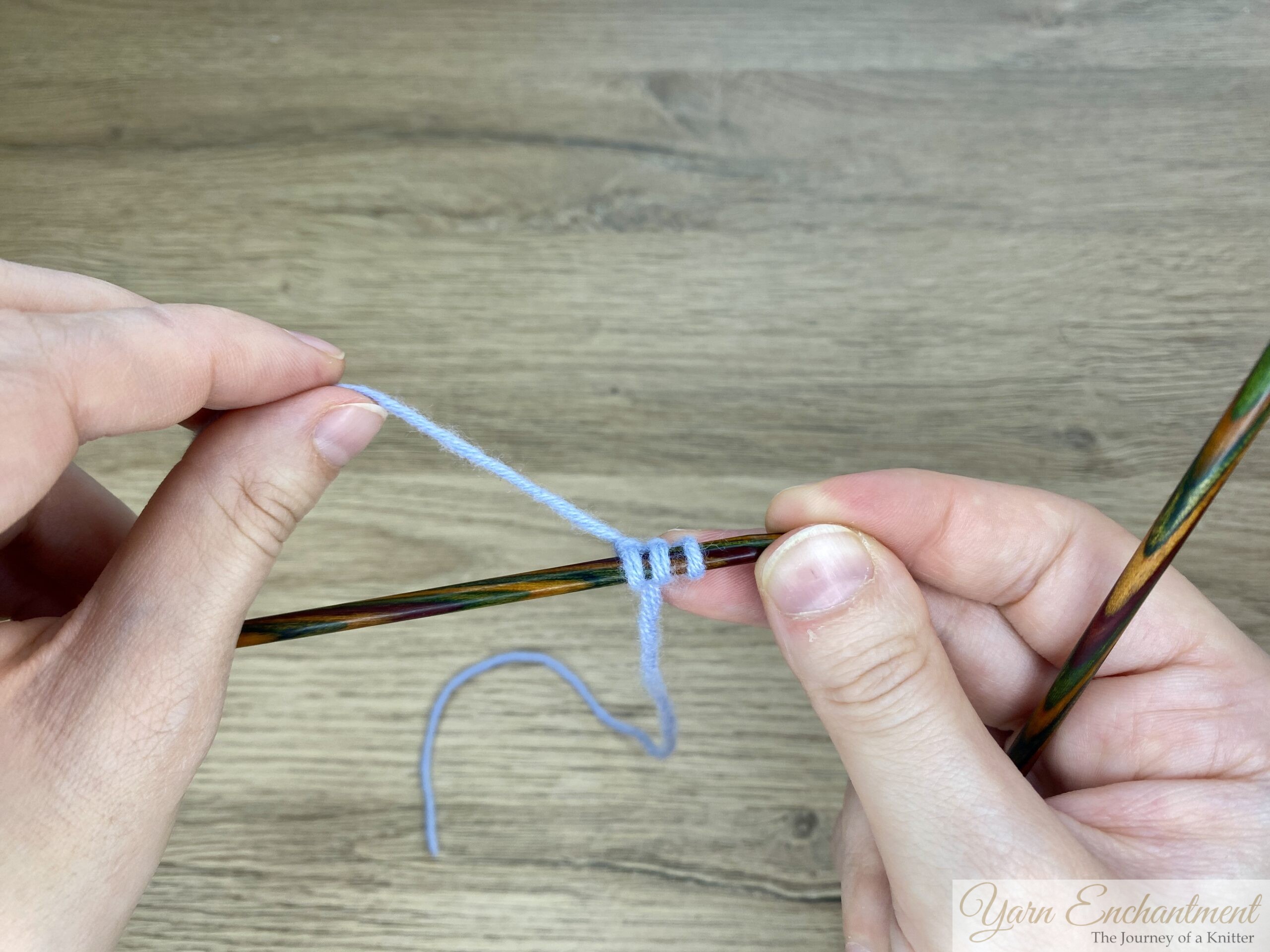 Two hands holding a wooden knitting needle with three light blue stitches; the left hand is preparing the working yarn to knit the next stitch.