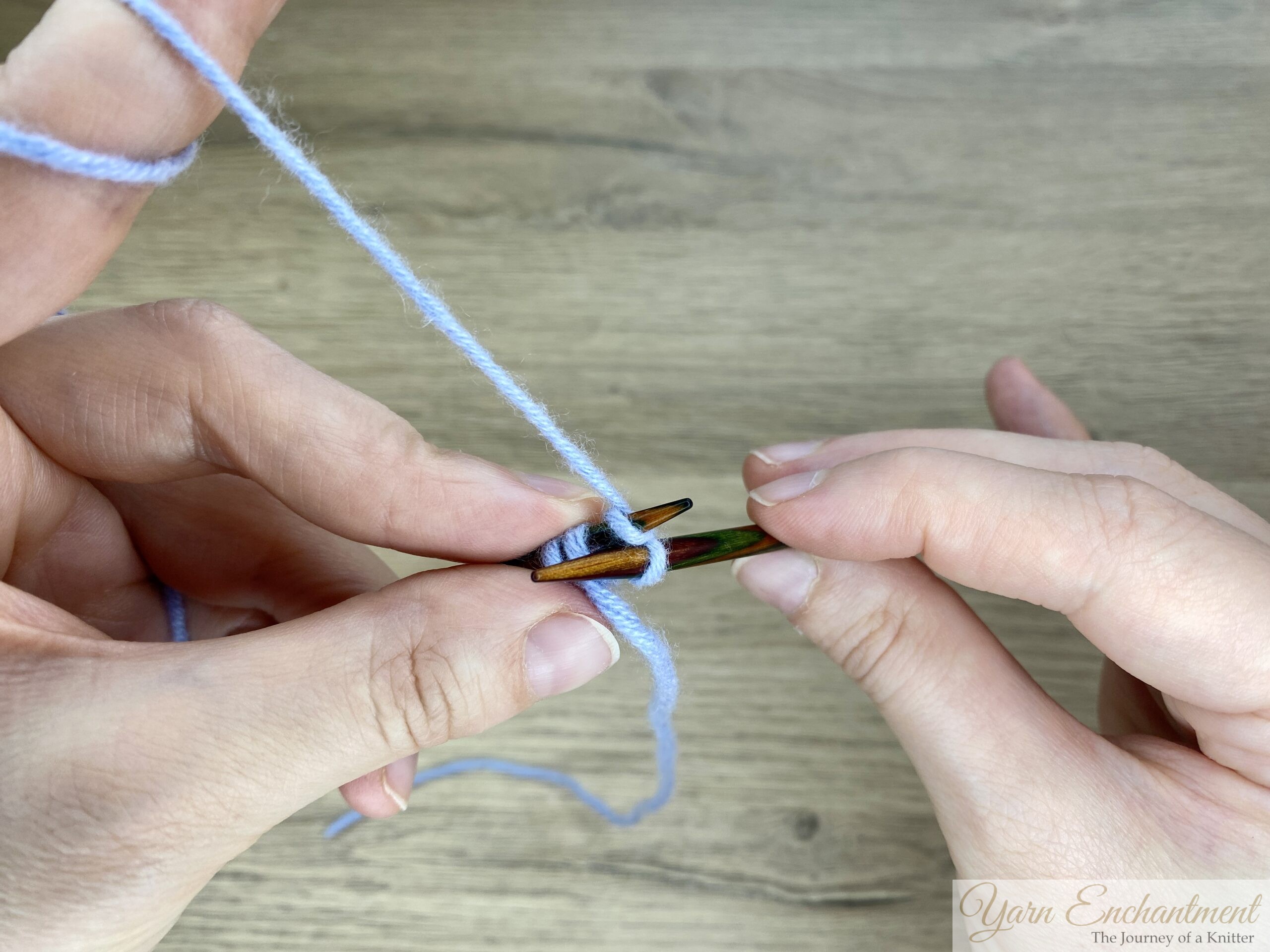 Hands holding a wooden needle, inserting the tip into the stitch and wrapping the yarn around, demonstrating the first knit stitch in the i-cord process.