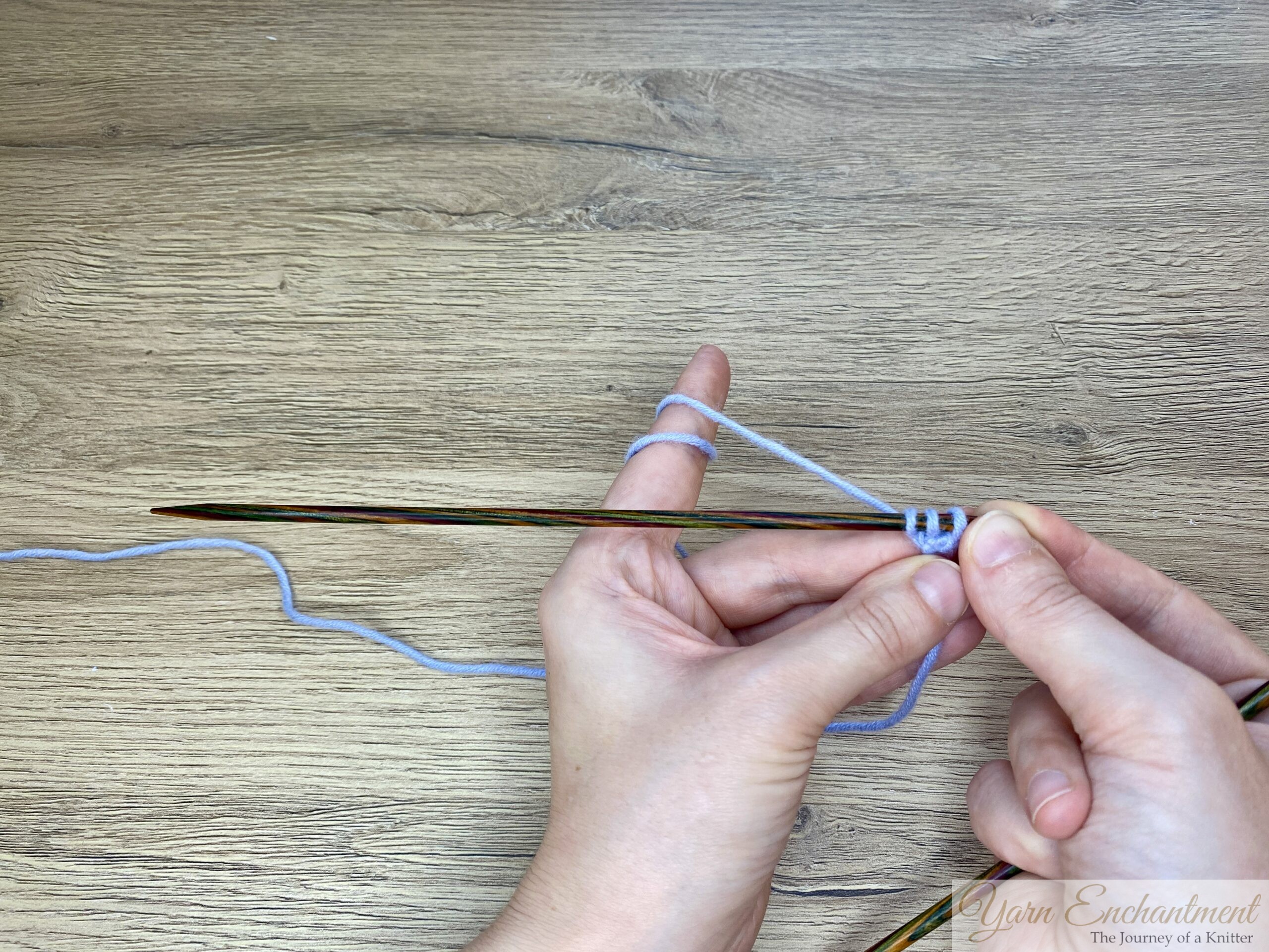 Both hands holding a wooden knitting needle with three stitches of light blue yarn, viewed from above, as the stitches are once again at the left end of the needle, ready for the next row in i-cord knitting.