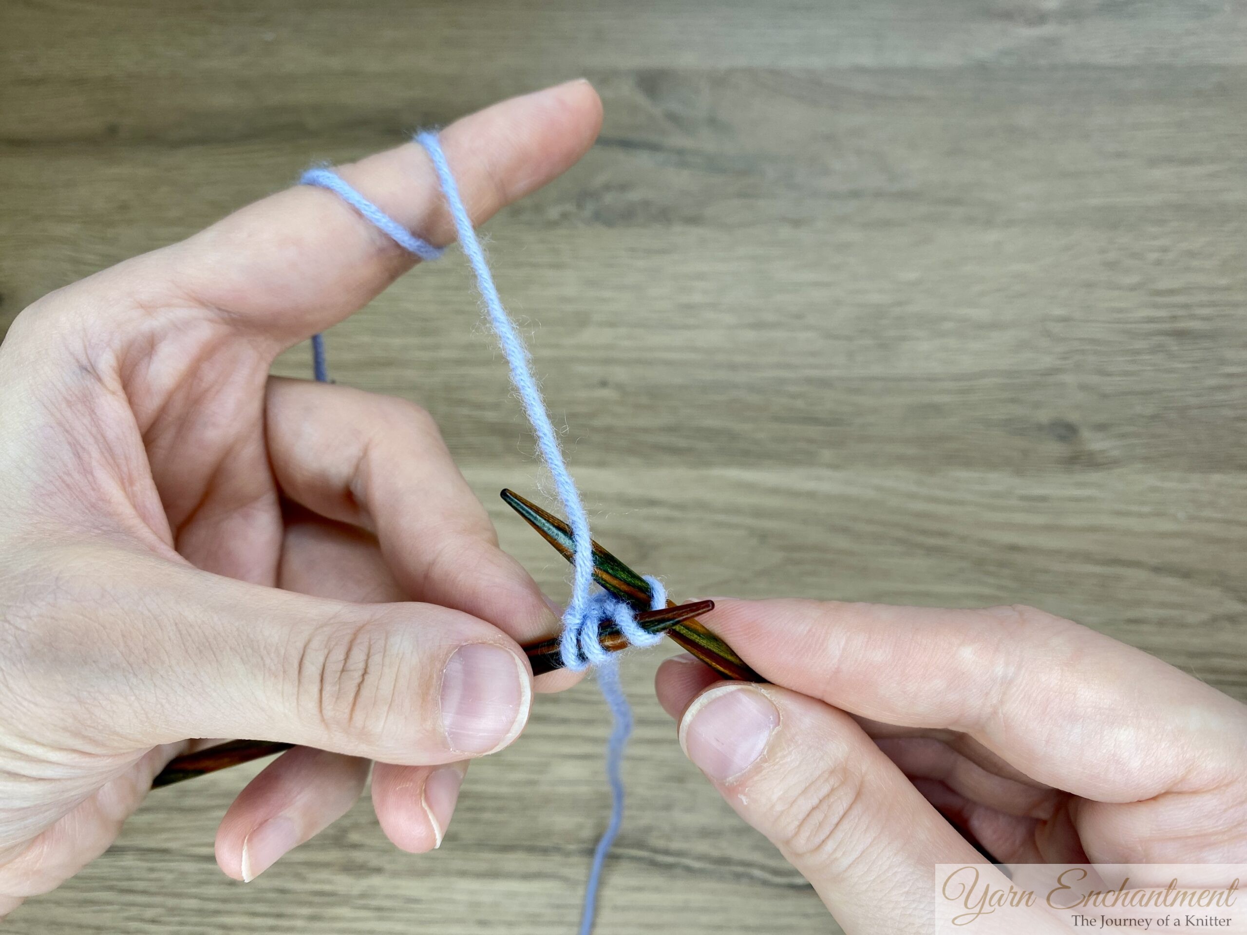 Two hands holding a wooden knitting needle with three stitches of light blue yarn; the working yarn is wrapped around the fingers, demonstrating the i-cord knitting technique.