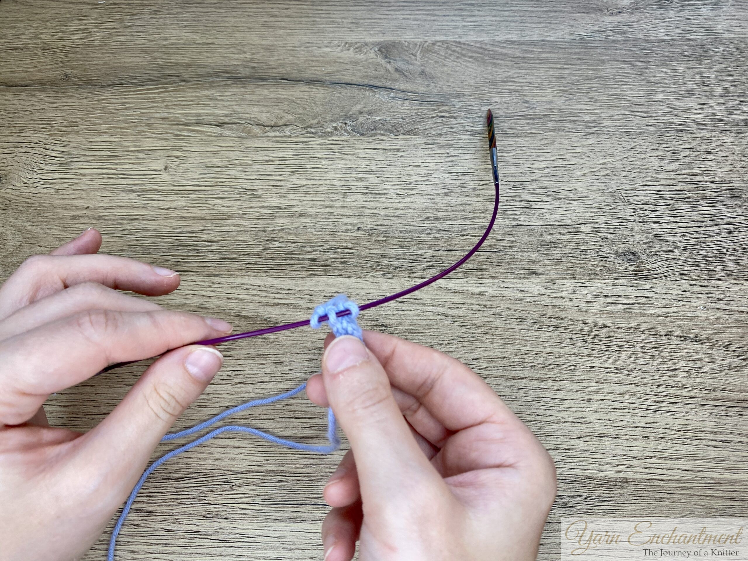 Hands holding a circular needle with blue I-cord stitches positioned on the cable, showing the stitches about to be slid over to the needle tip.