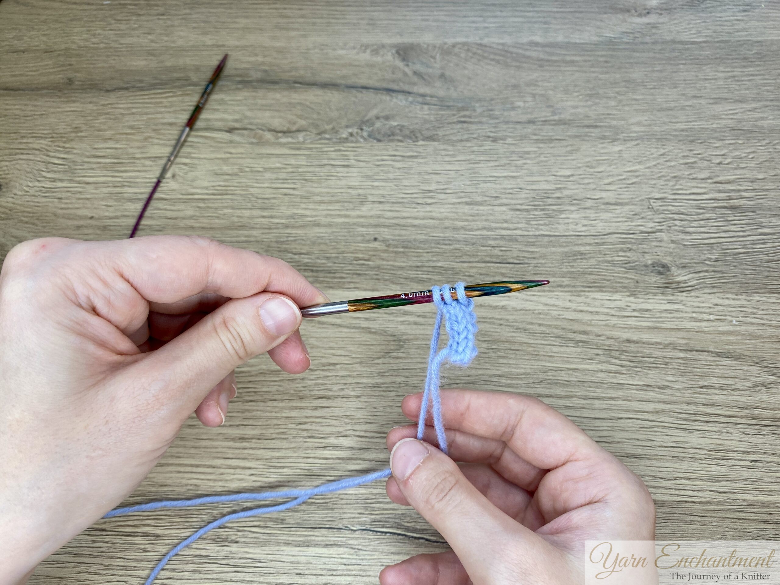 Both hands display the circular needle with completed blue I-cord; all stitches are now collected on the needle tip, illustrating the finished step.