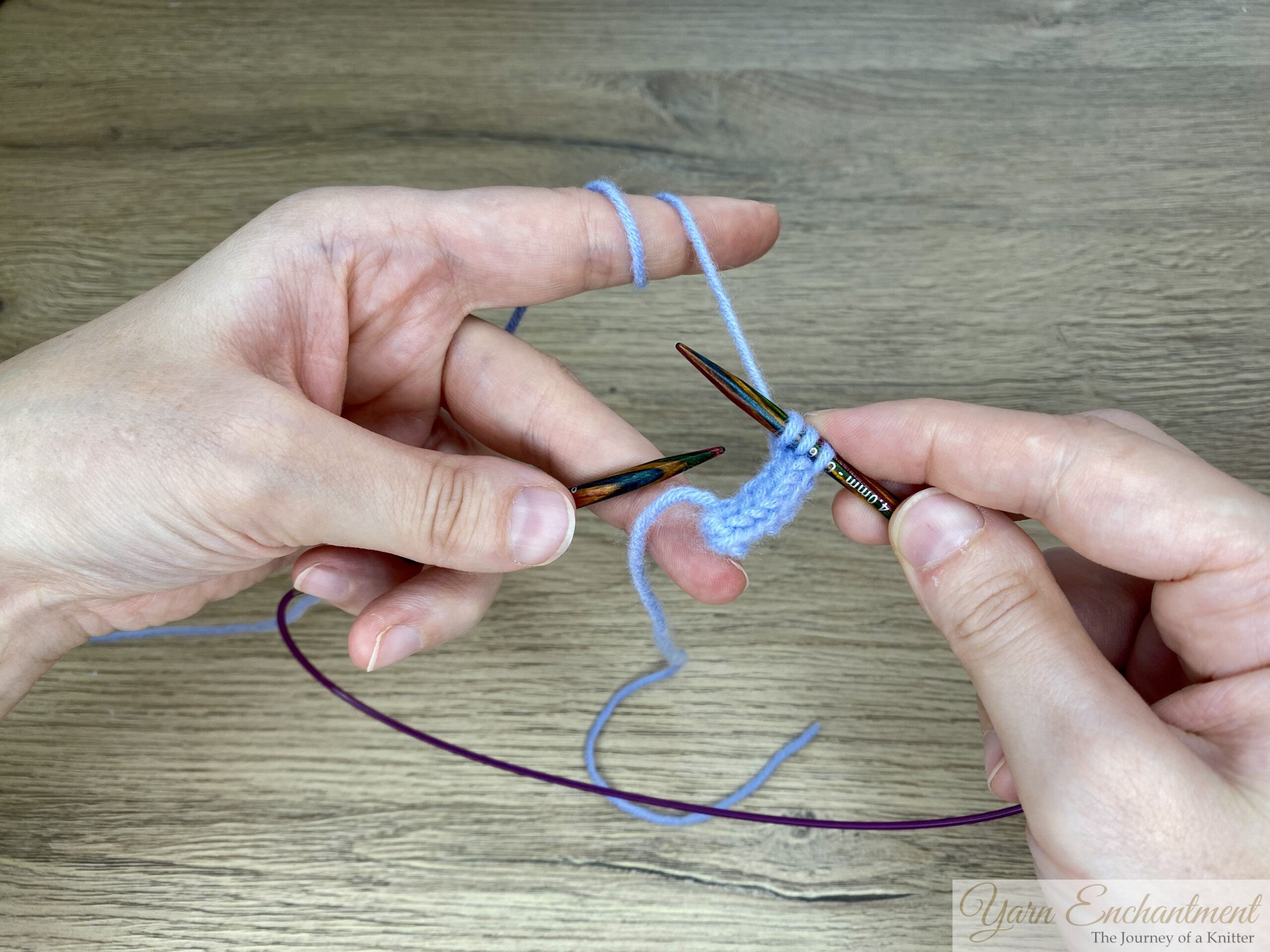 Hands holding circular knitting needles with blue yarn after sliding the I-cord stitches to the other side of the needle, preparing for the next row.