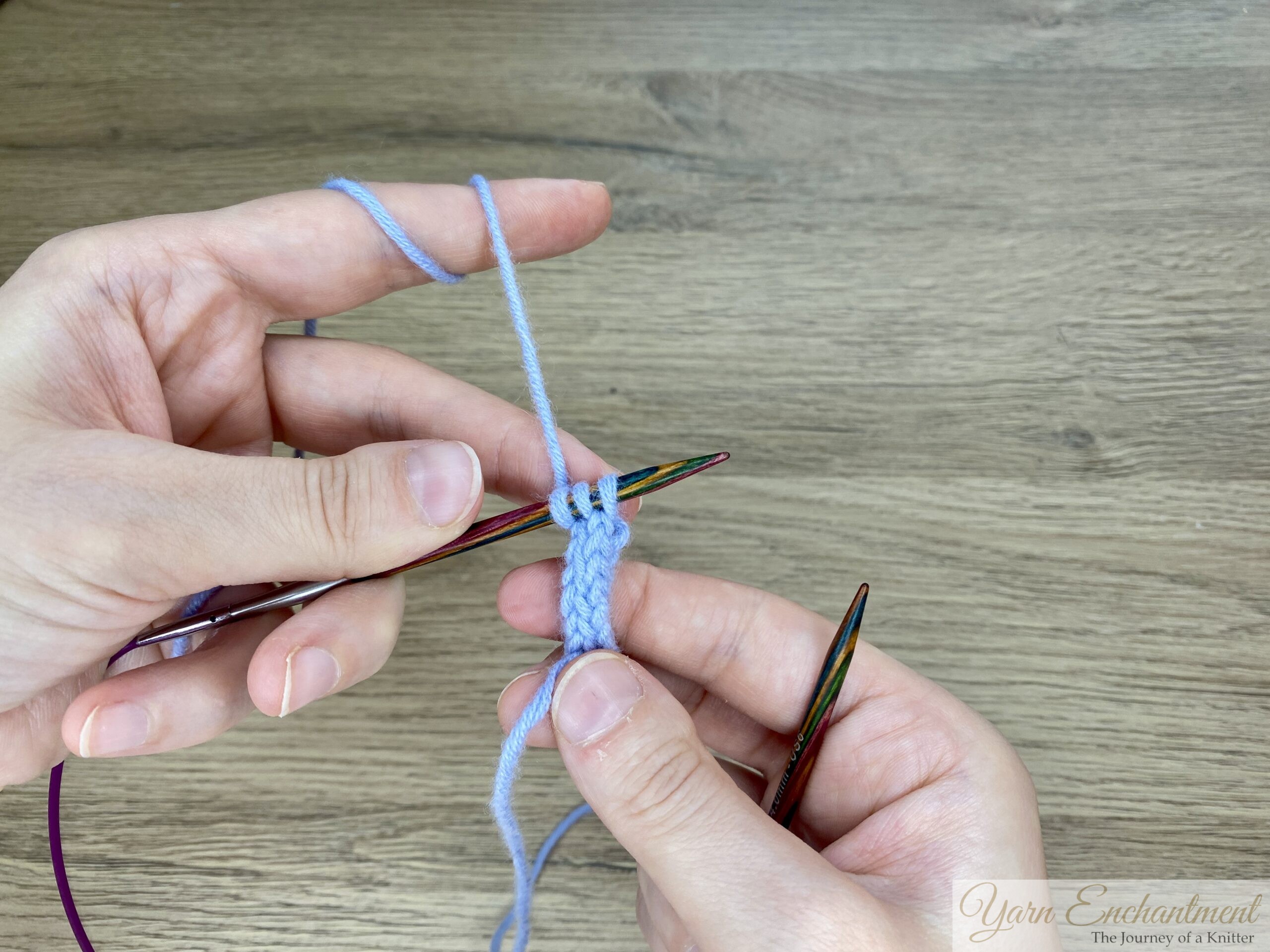 Knitted I-cord in progress on circular knitting needles, with three stitches visible and yarn tensioned over the left hand.