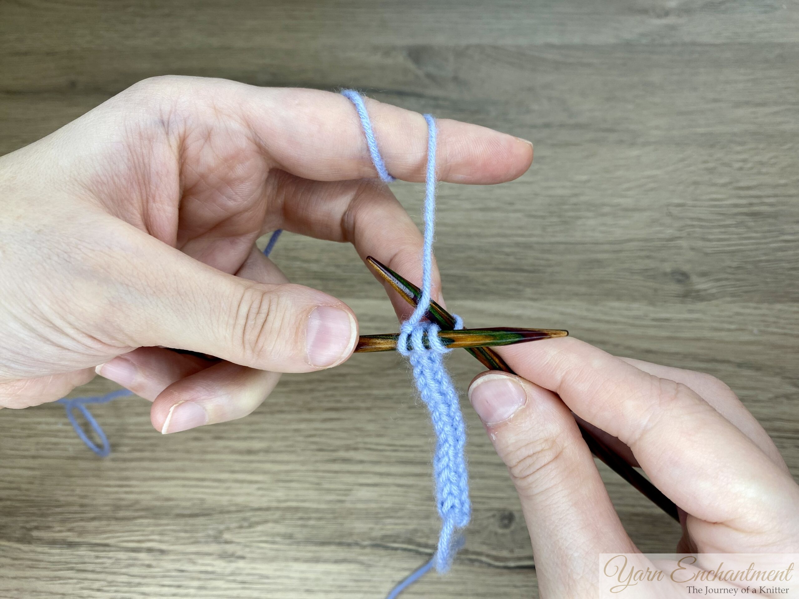 Close-up of hands binding off the end of a blue I-cord, with one stitch remaining on the right needle, demonstrating the finishing step.