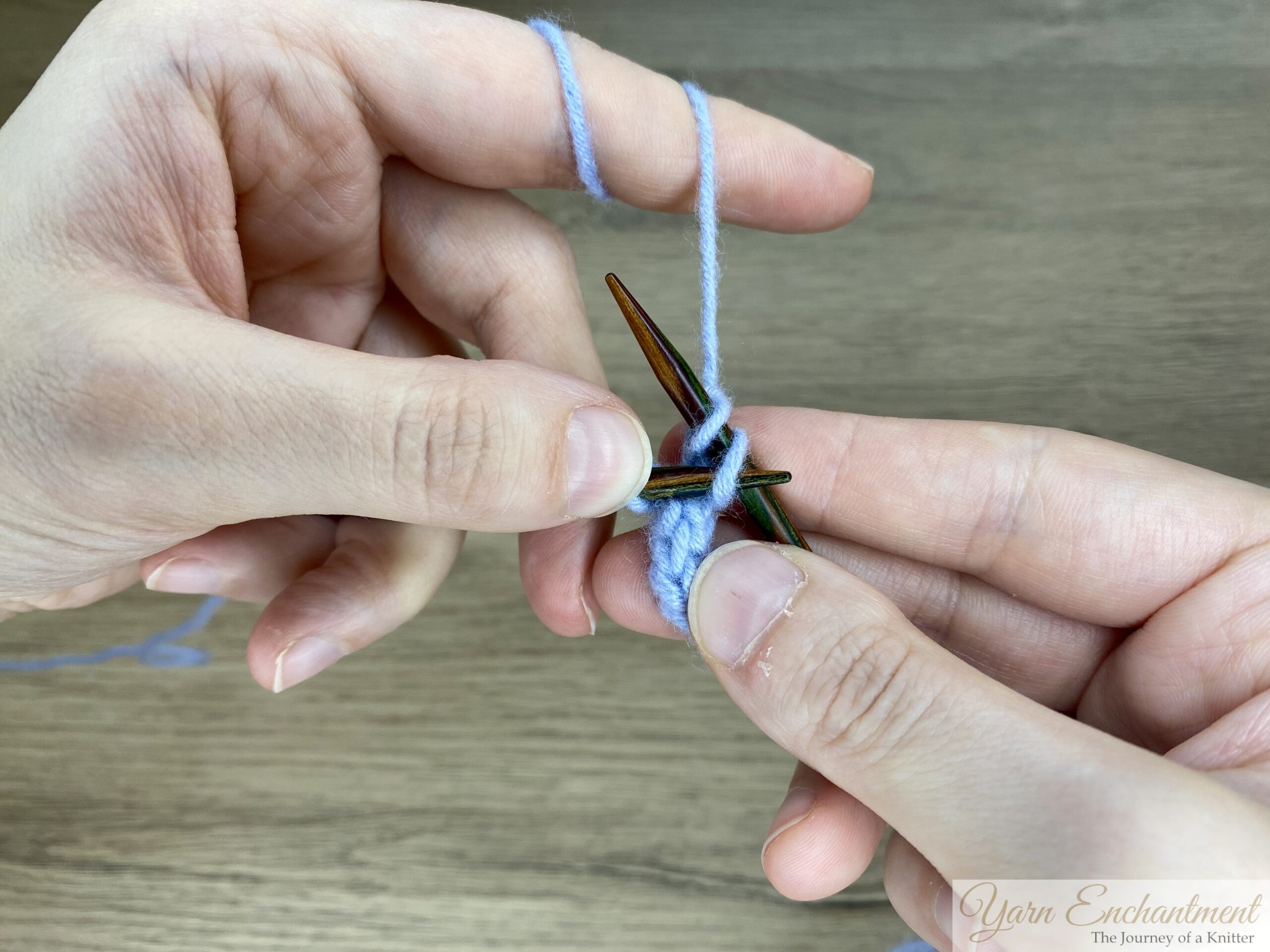 Hands holding knitting needles and binding off the final stitches of a blue I-cord, showing the last loops ready to be secured.