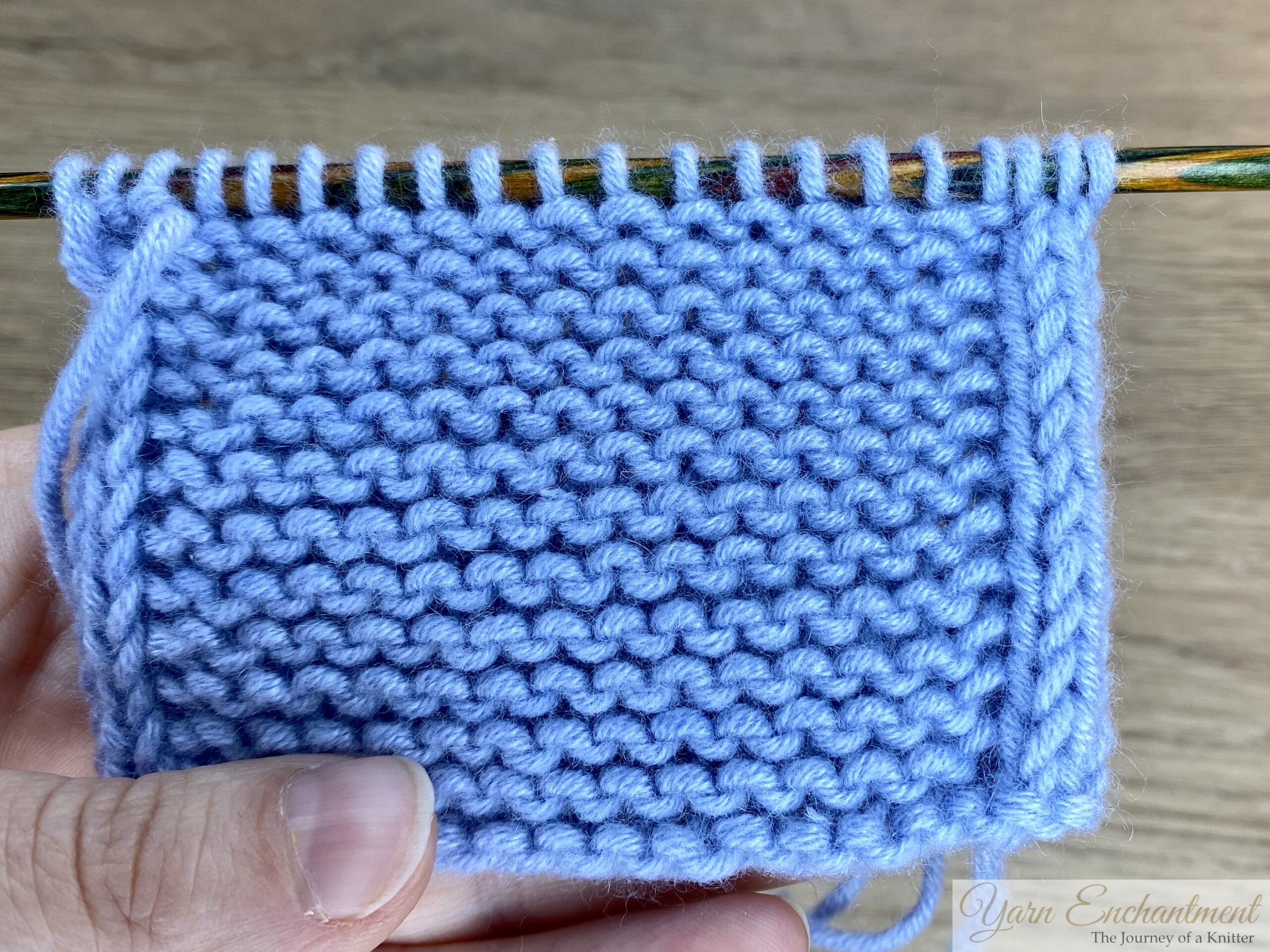 Swatch of blue garter stitch knitting on a wooden needle, featuring neat I-cord edging along both sides for a clean, finished border.