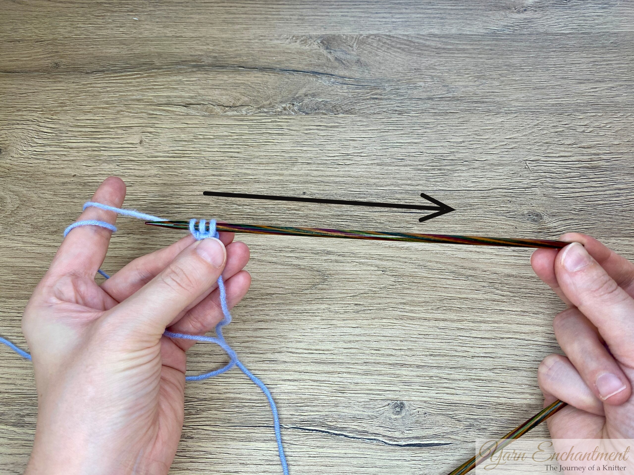 A pair of hands holding a wooden knitting needle with three stitches of light blue yarn, with an added black arrow drawn above the needle, showing the direction to slide the stitches to the right.