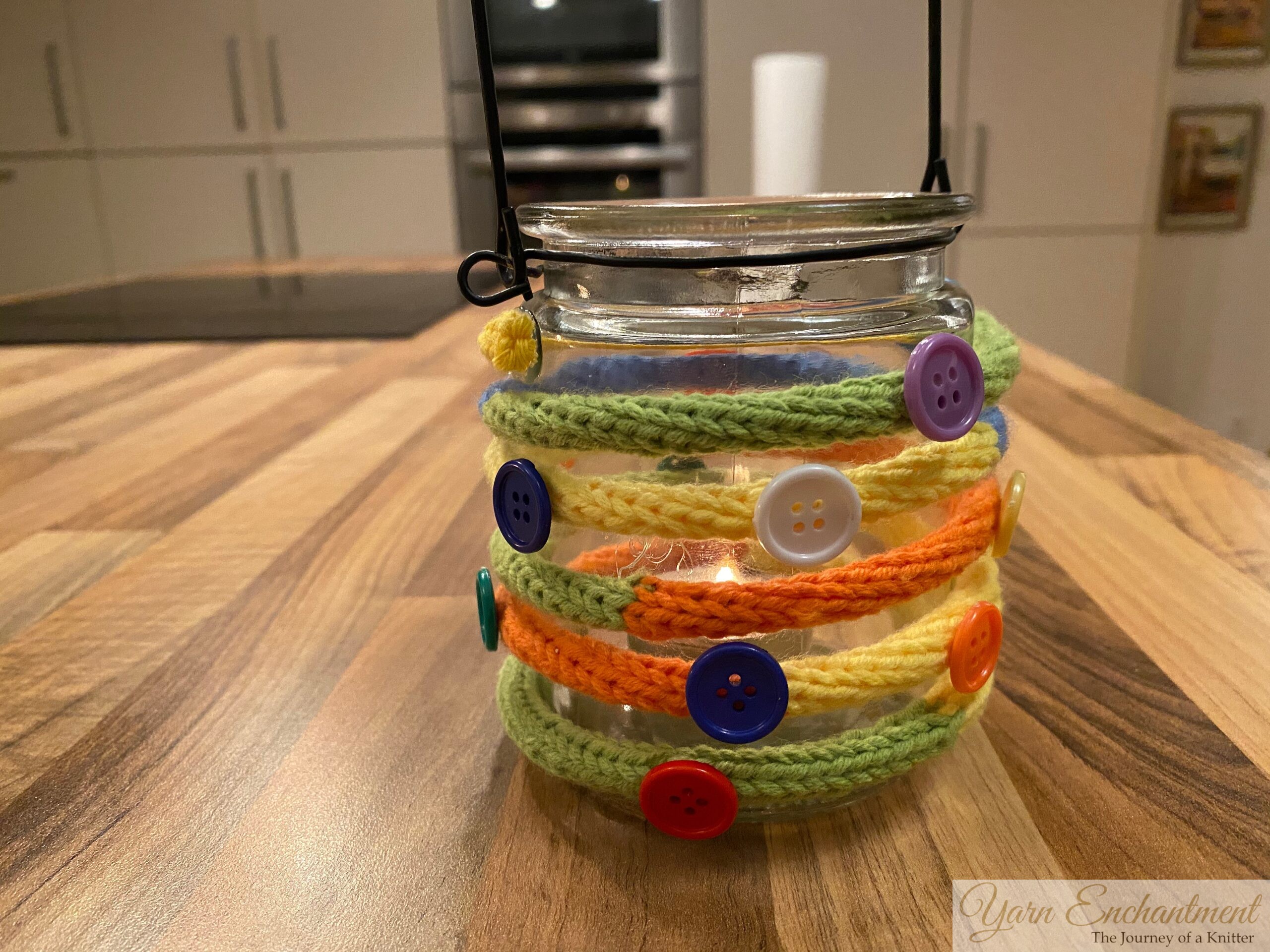 HGlass jar wrapped with colorful knitted I-cords and decorated with various buttons, displayed on a wooden kitchen counter.