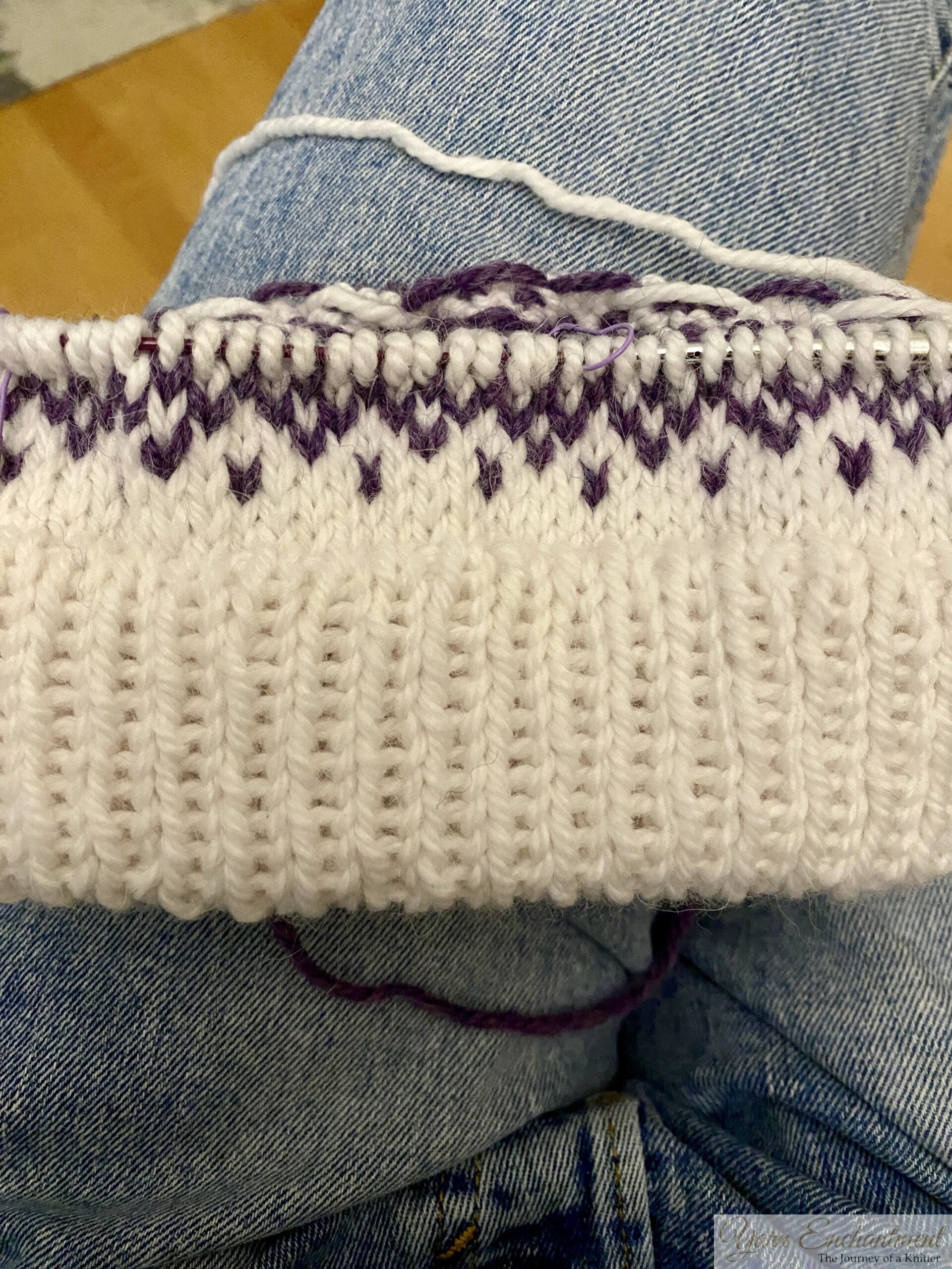 White knitted beanie with a deep double-folded ribbed brim and a purple swirling colorwork band, styled flat on a wooden background with pinecones and green leaves.