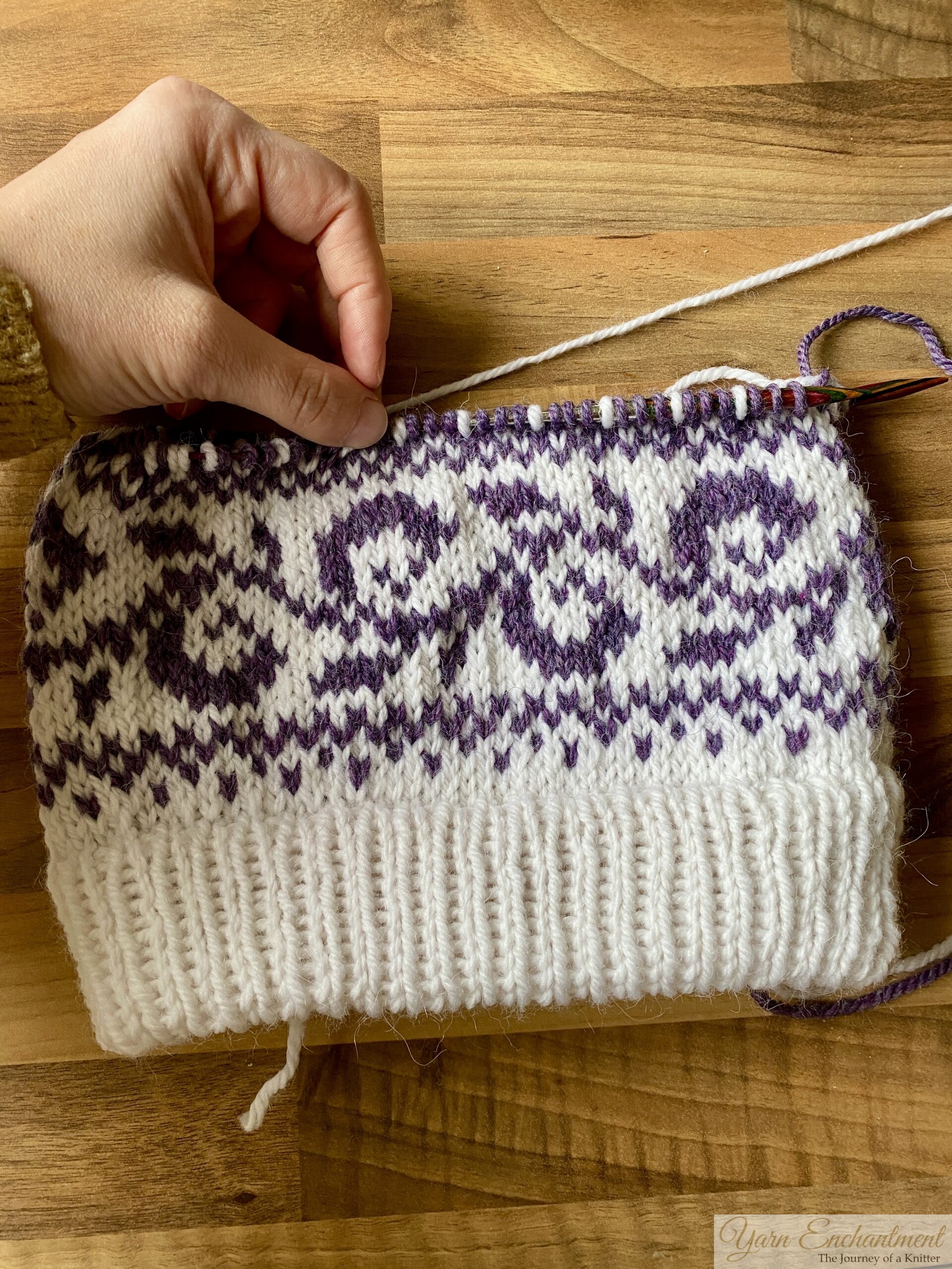 White knitted beanie with a deep double-folded ribbed brim and a purple swirling colorwork band, styled flat on a wooden background with pinecones and green leaves.