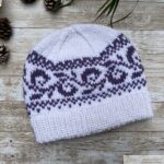 White knitted beanie with a deep double-folded ribbed brim and a purple swirling colorwork band, styled flat on a wooden background with pinecones and green leaves.
