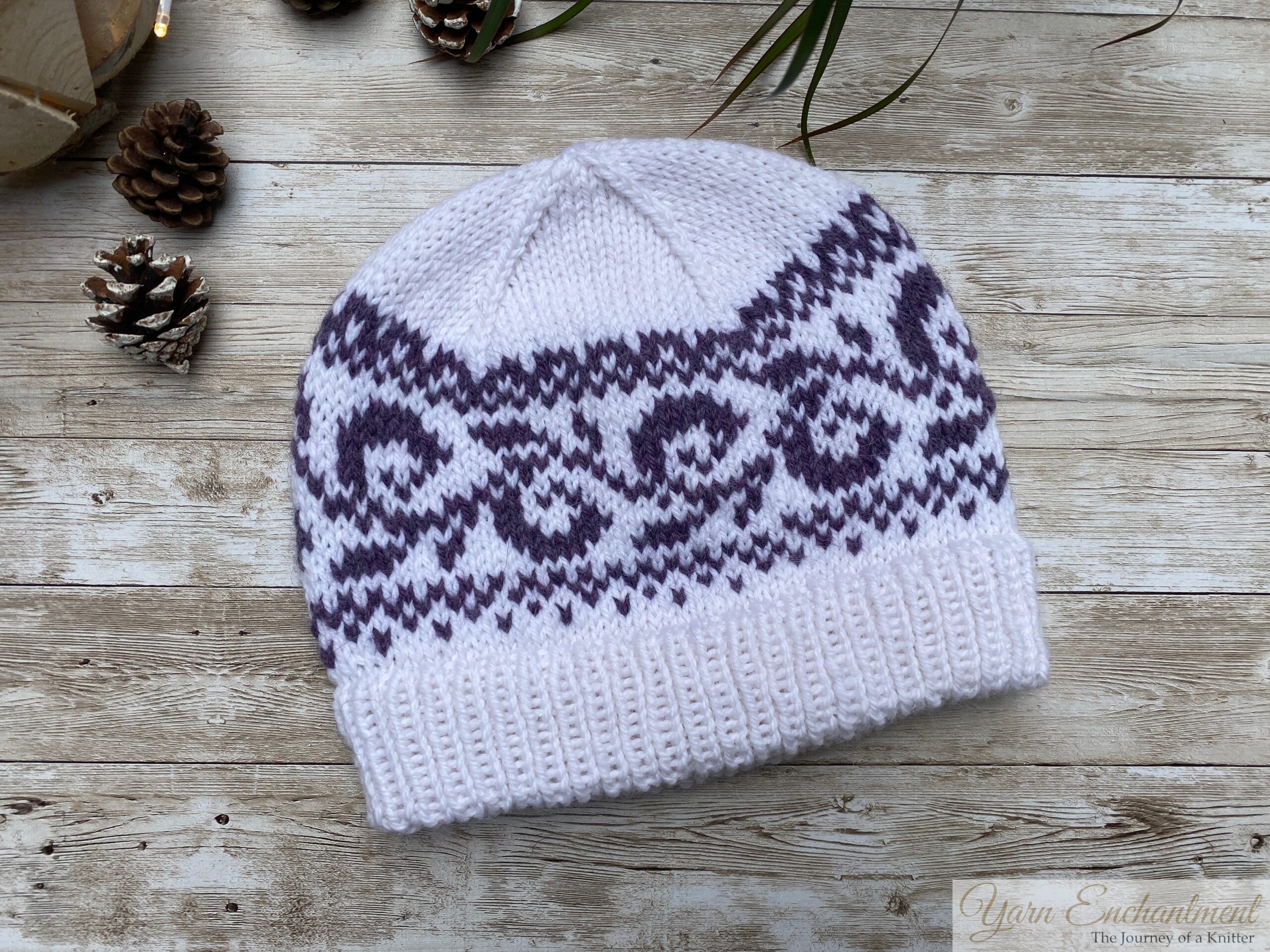 White knitted beanie with a deep double-folded ribbed brim and a purple swirling colorwork band, styled flat on a wooden background with pinecones and green leaves.