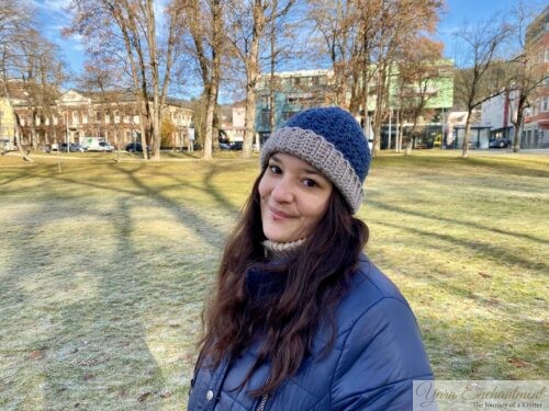 Person wearing a blue crochet star stitch hat with a beige brim and a navy winter coat, standing in a frosty city park with blurred face for privacy.