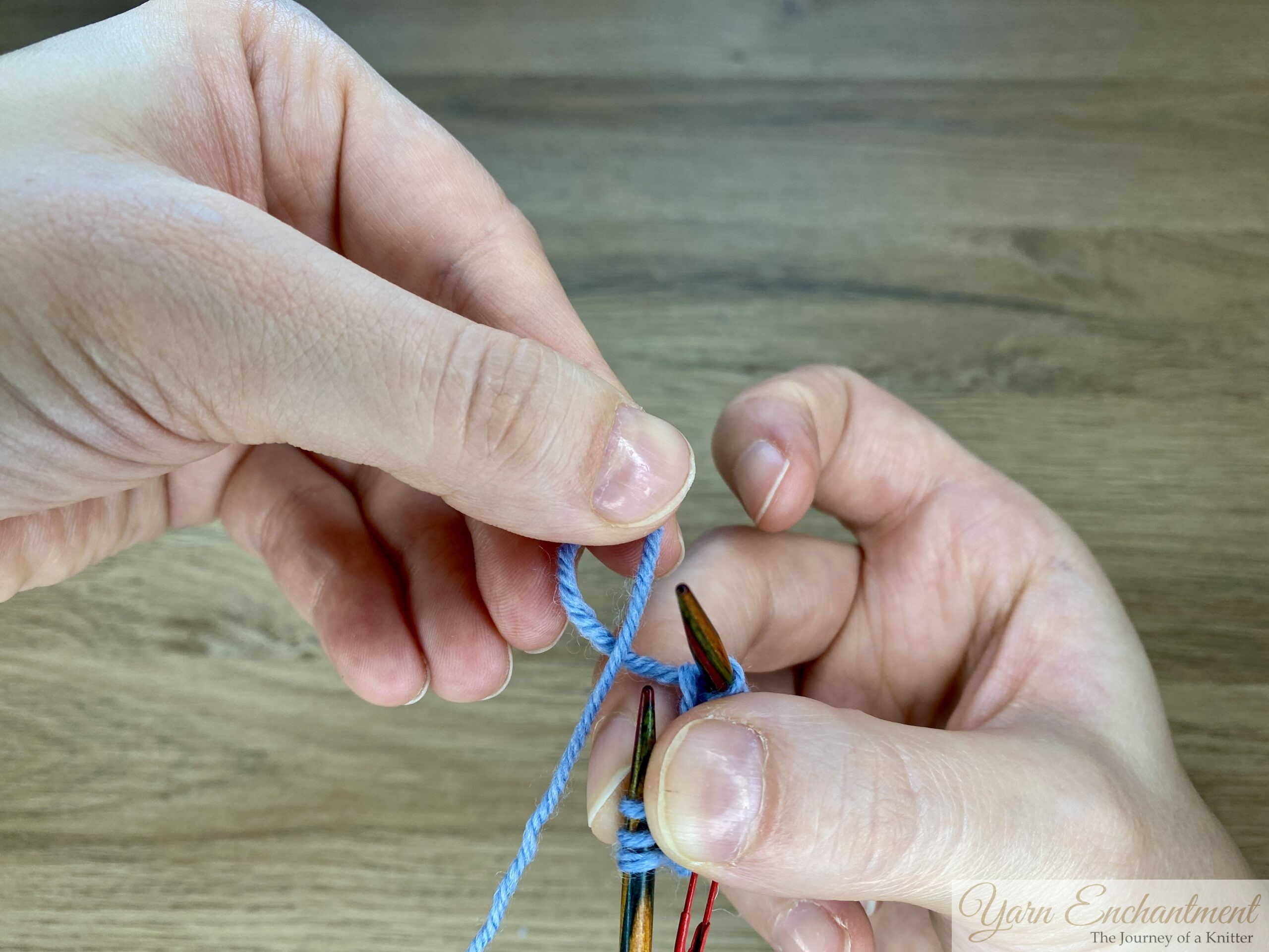 Hands holding blue yarn and forming a backward loop around the needle to work the first increase between the I-cord edges.