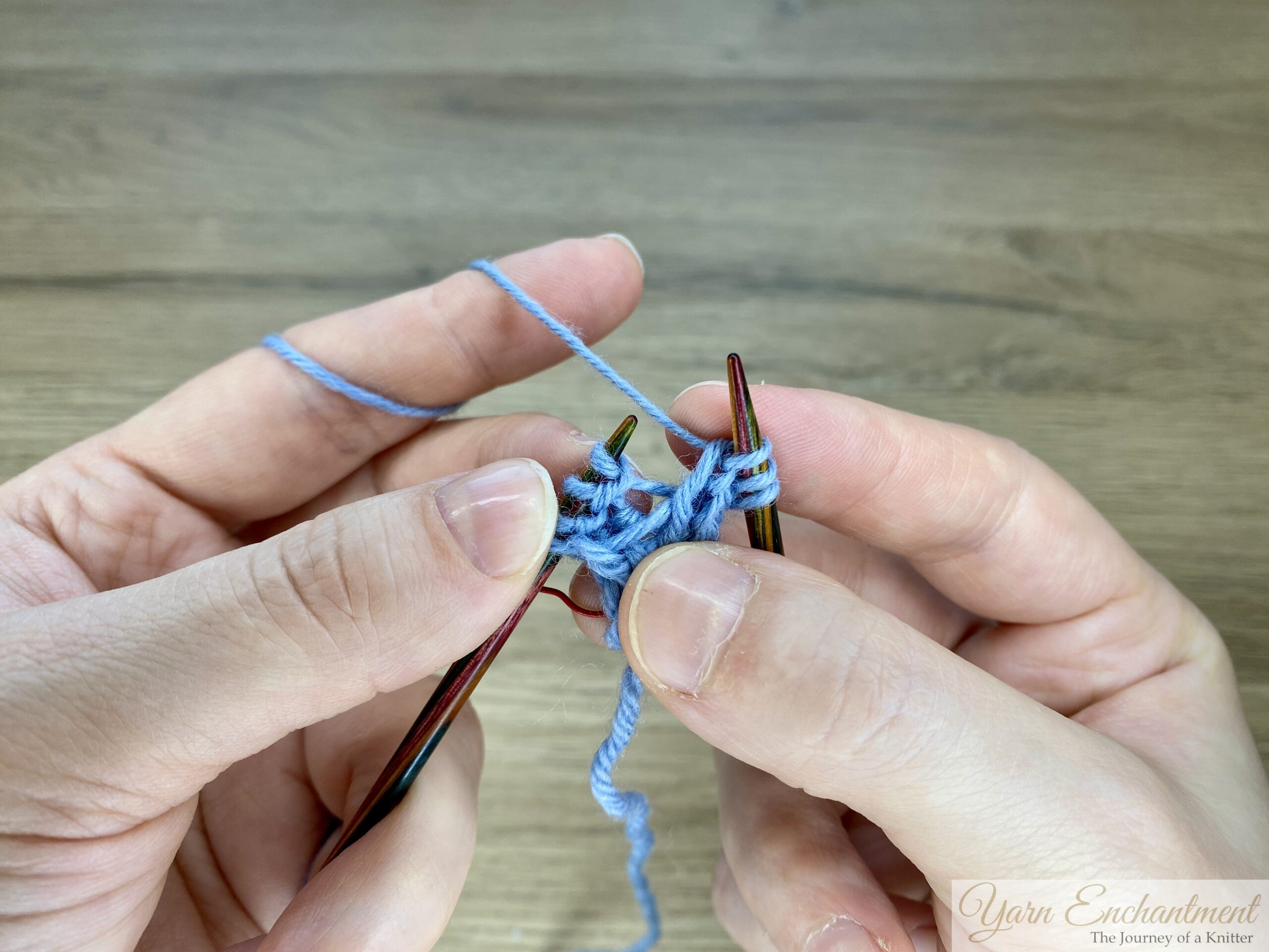 Blue garter-stitch swatch with I-cord edging on wooden needles showing the wrong side after the increase