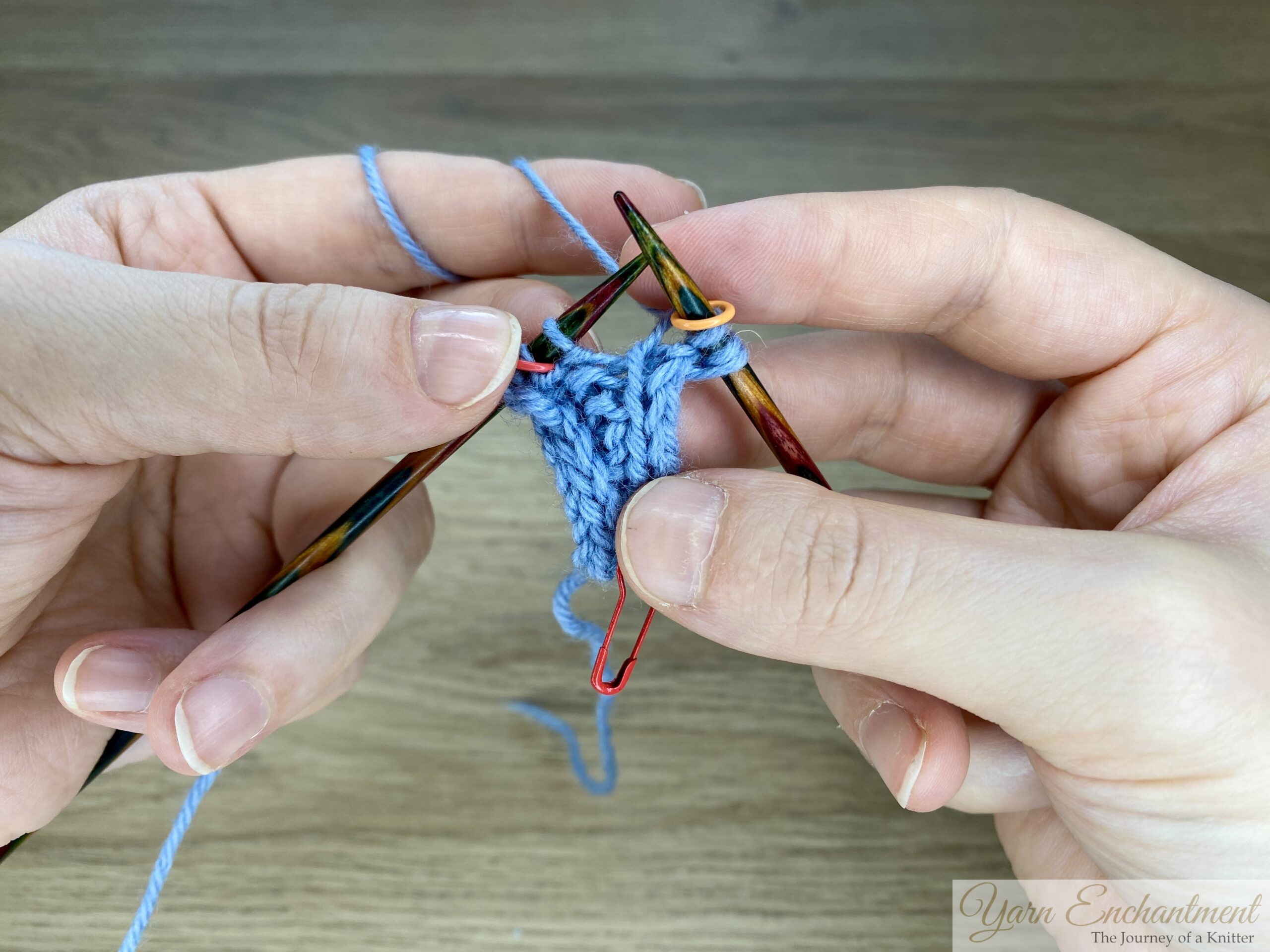 Hands knitting a blue Sophie-style swatch, working a kfb increase just after the I-cord edge on multicoloured wooden needles