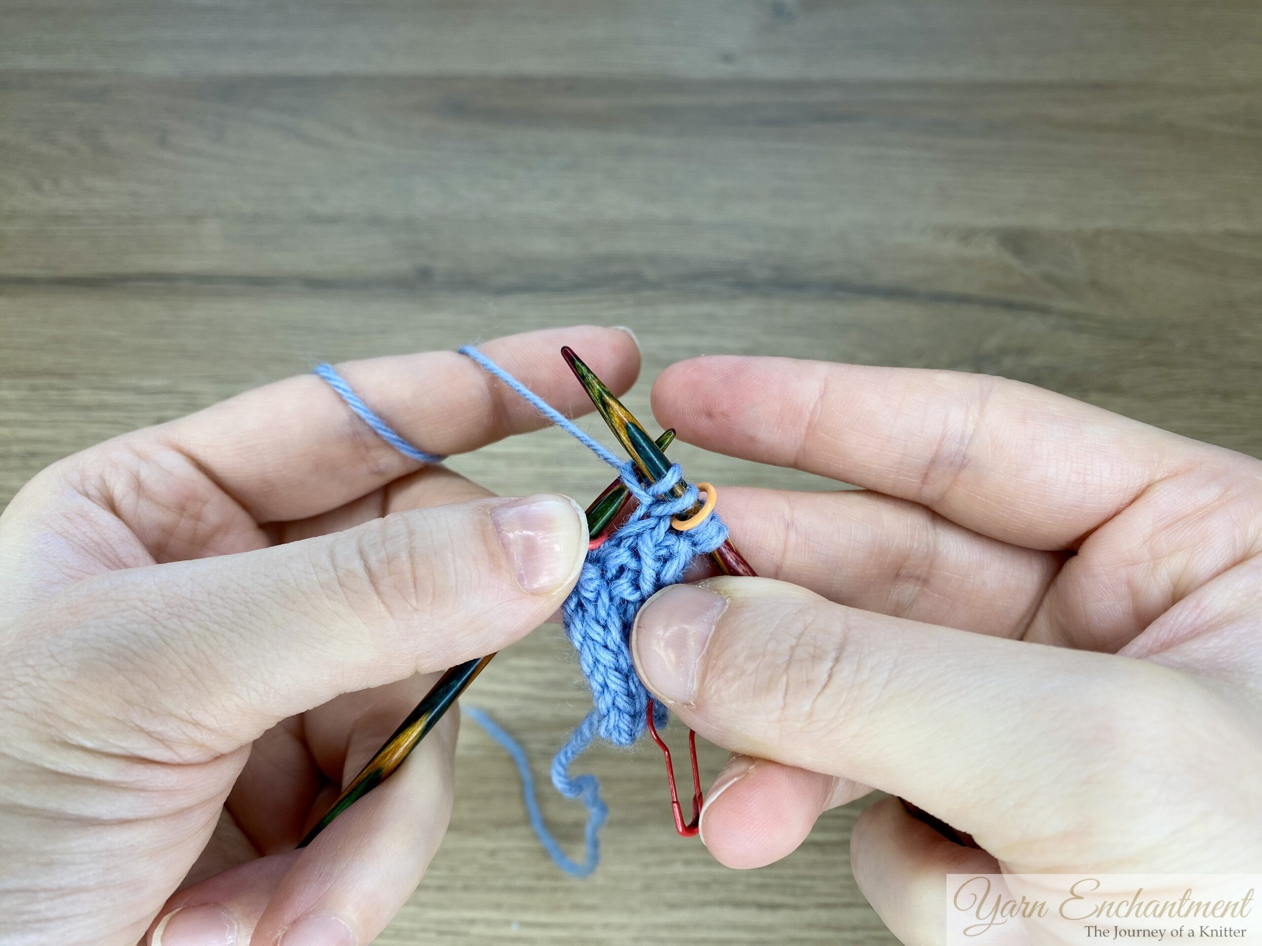 Close-up of a knitter inserting the right needle into the first stitch after the I-cord edge to work a knit front and back increase on a blue garter-stitch swatch