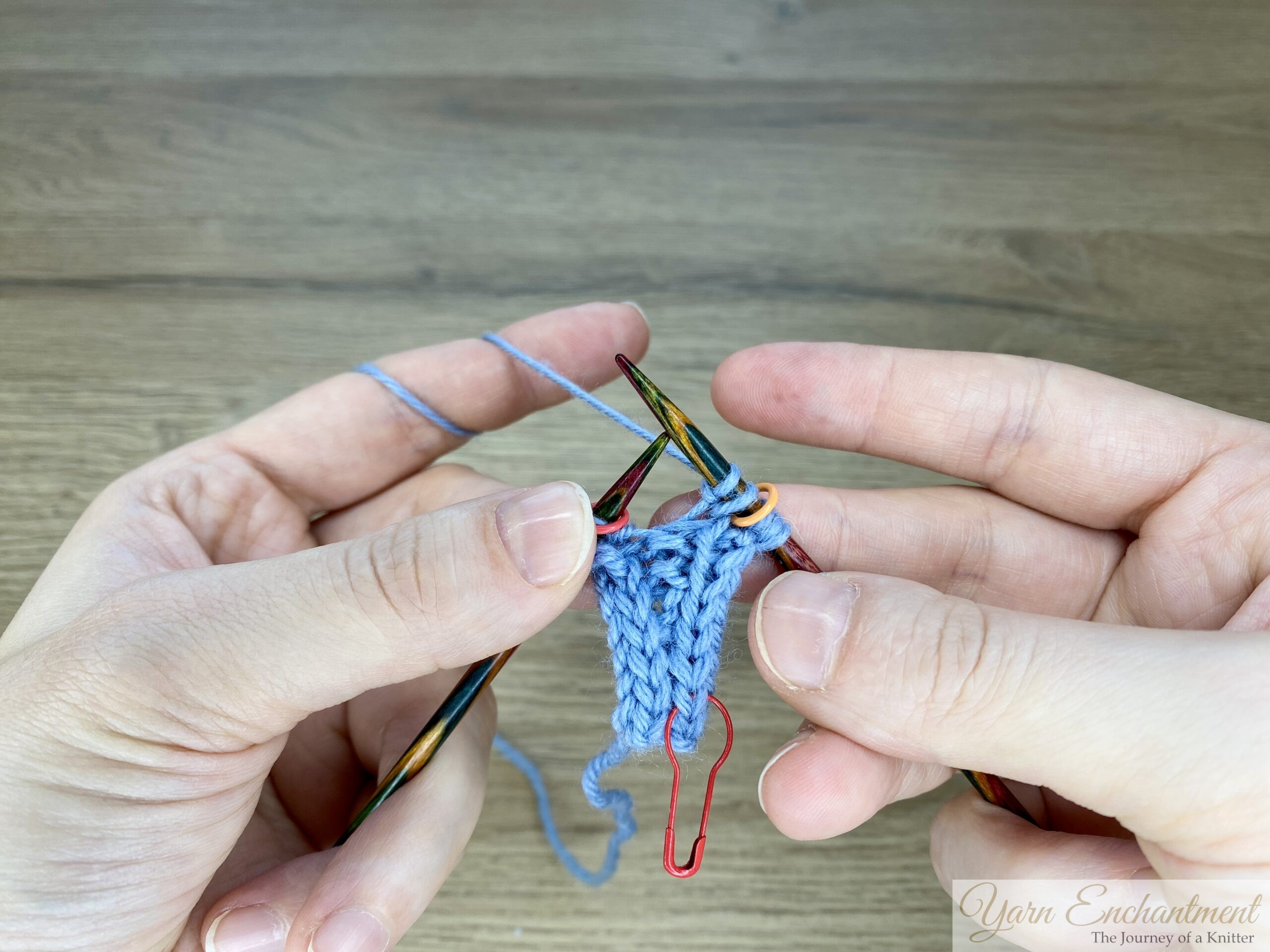 Blue garter-stitch swatch with I-cord edging on wooden needles, showing the completed kfb increase sitting neatly inside the edge stitch
