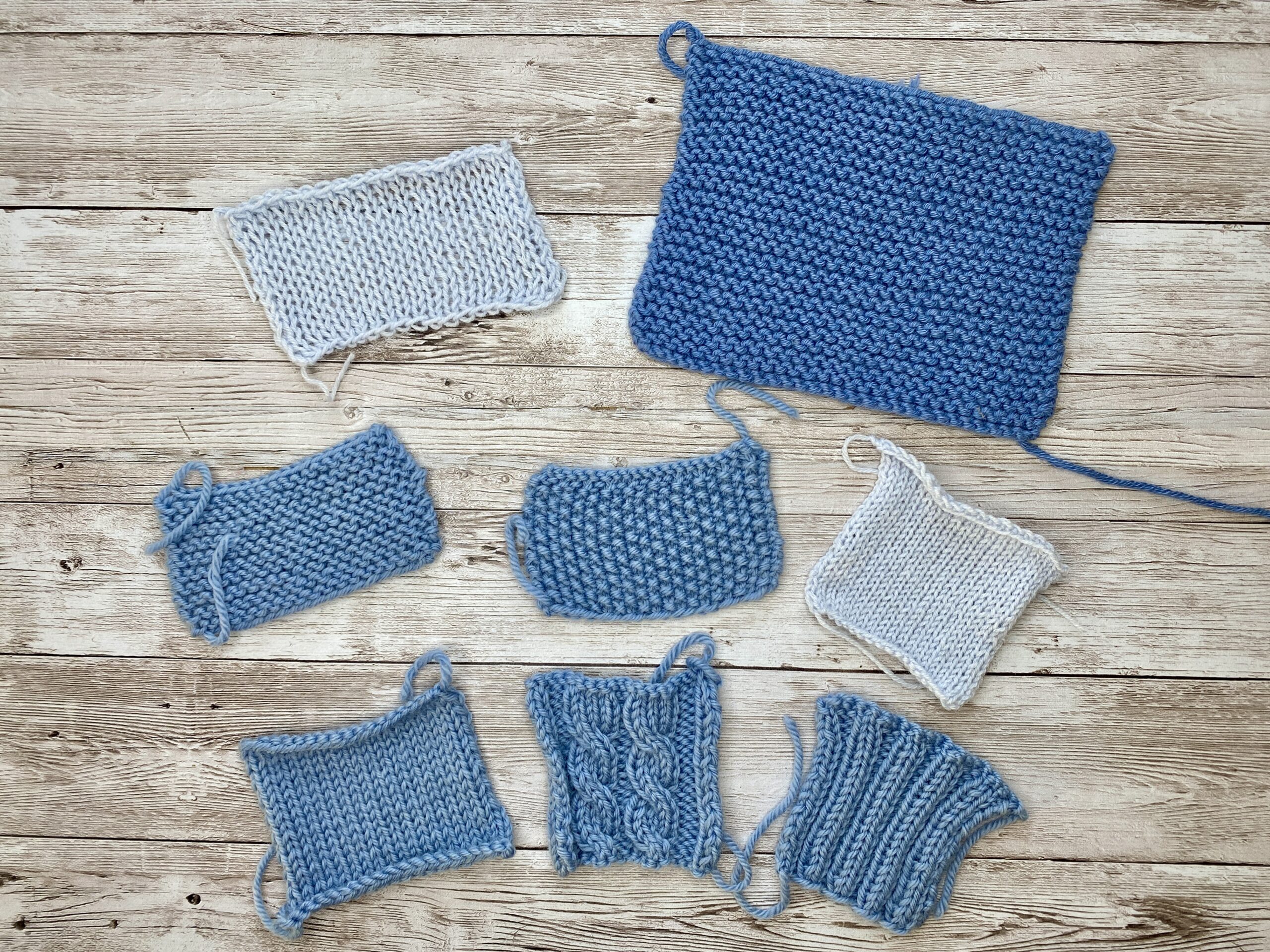 Several blue and light grey knitting gauge swatches in different stitch patterns and sizes laid out on a wooden background.