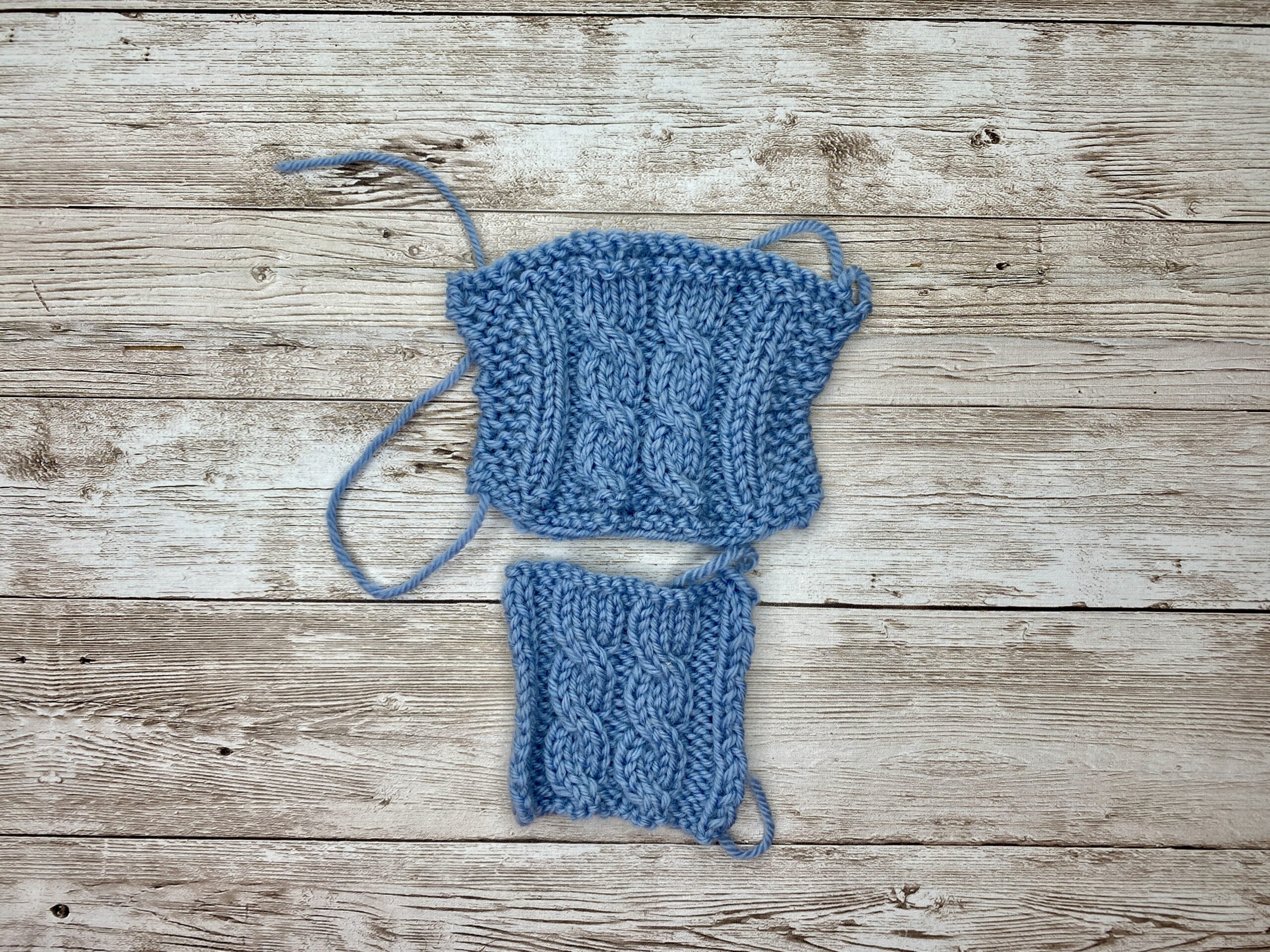 Blue cabled gauge swatch with garter borders above a borderless cabled swatch, both in the same yarn.