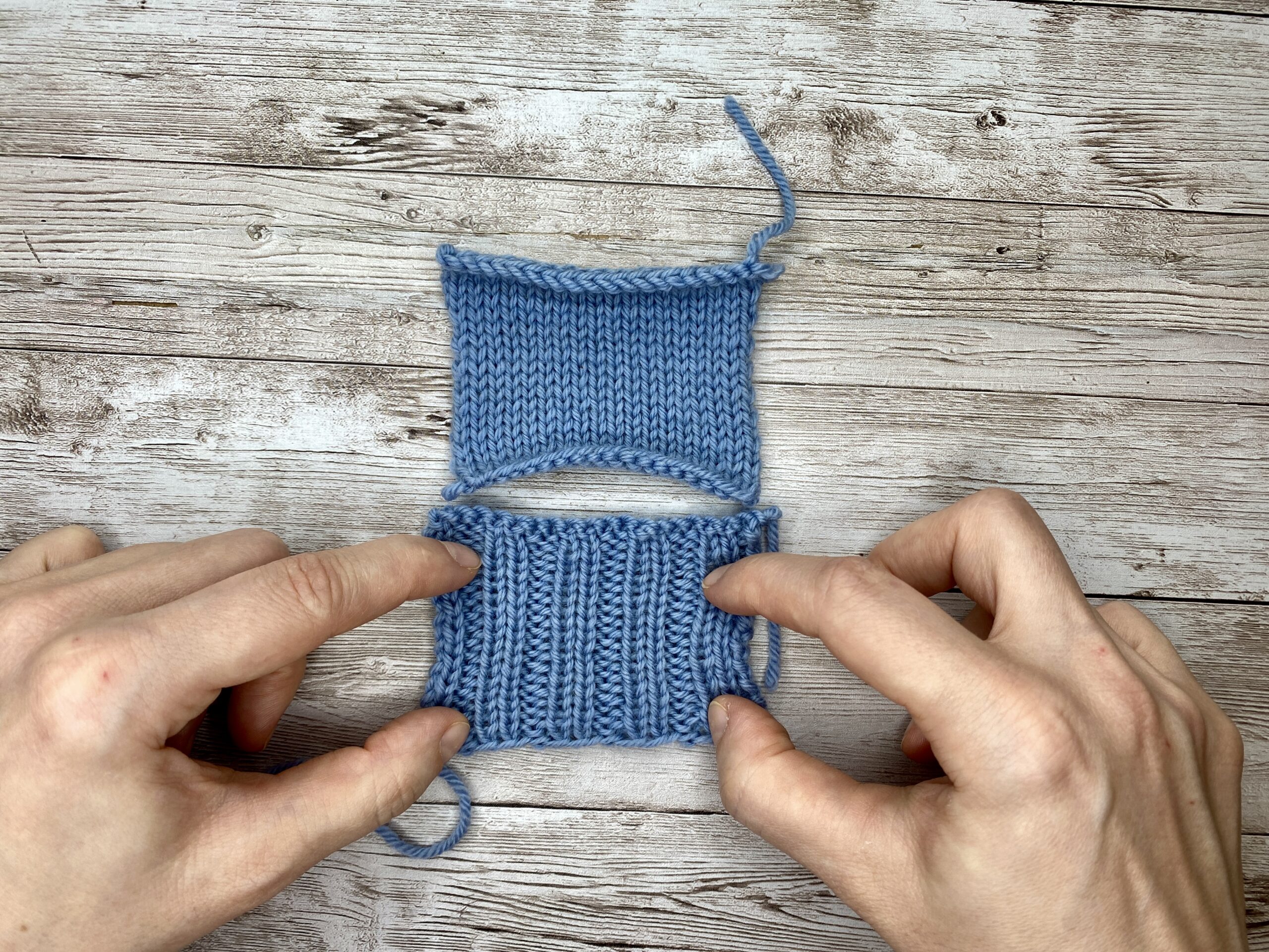 Hands holding a blue gauge swatch with garter borders, gently stretching the centre fabric.