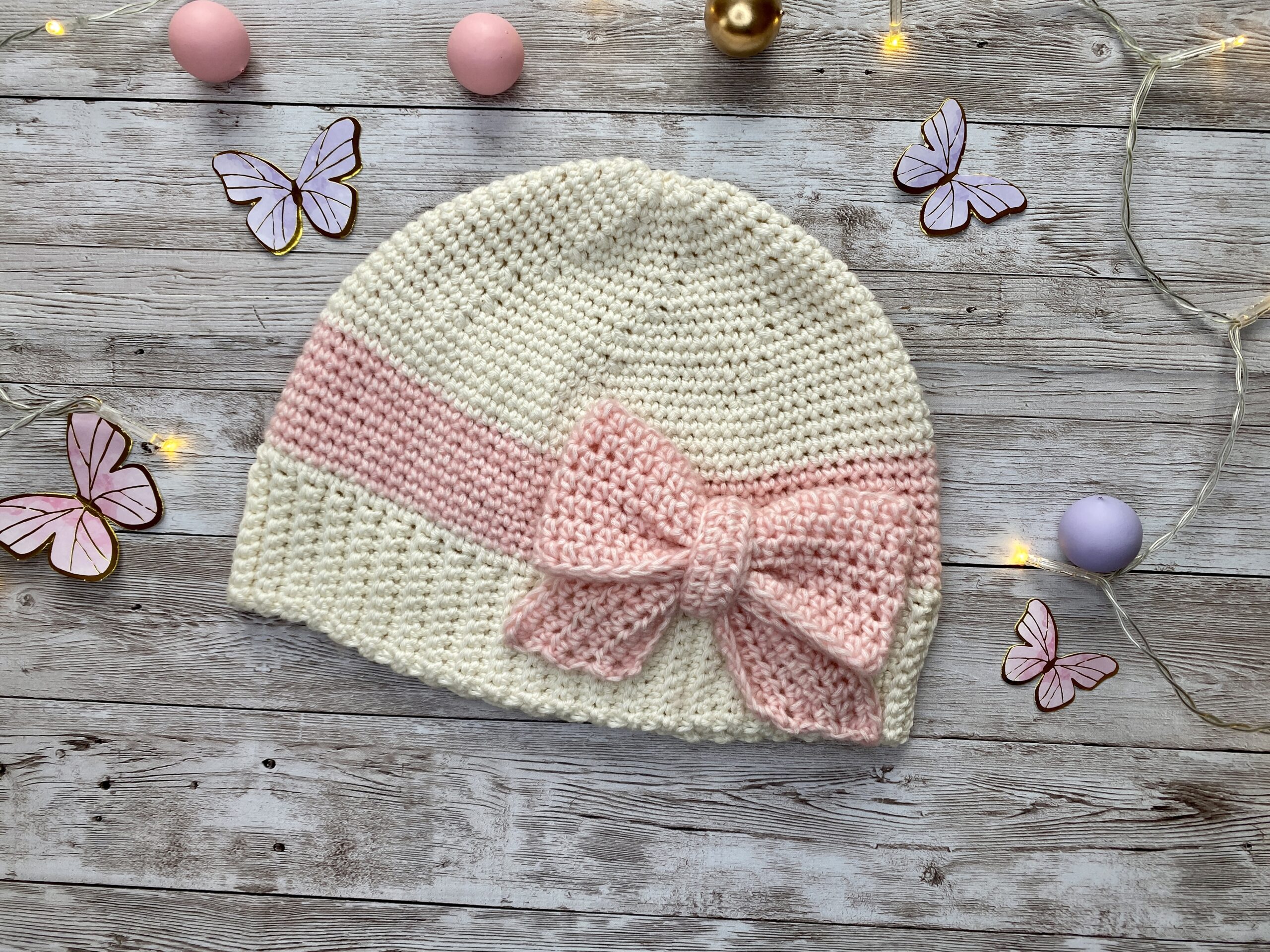 Flat lay of a handmade cream and blush crochet hat with a large pink bow, styled with soft decorative elements in the background.