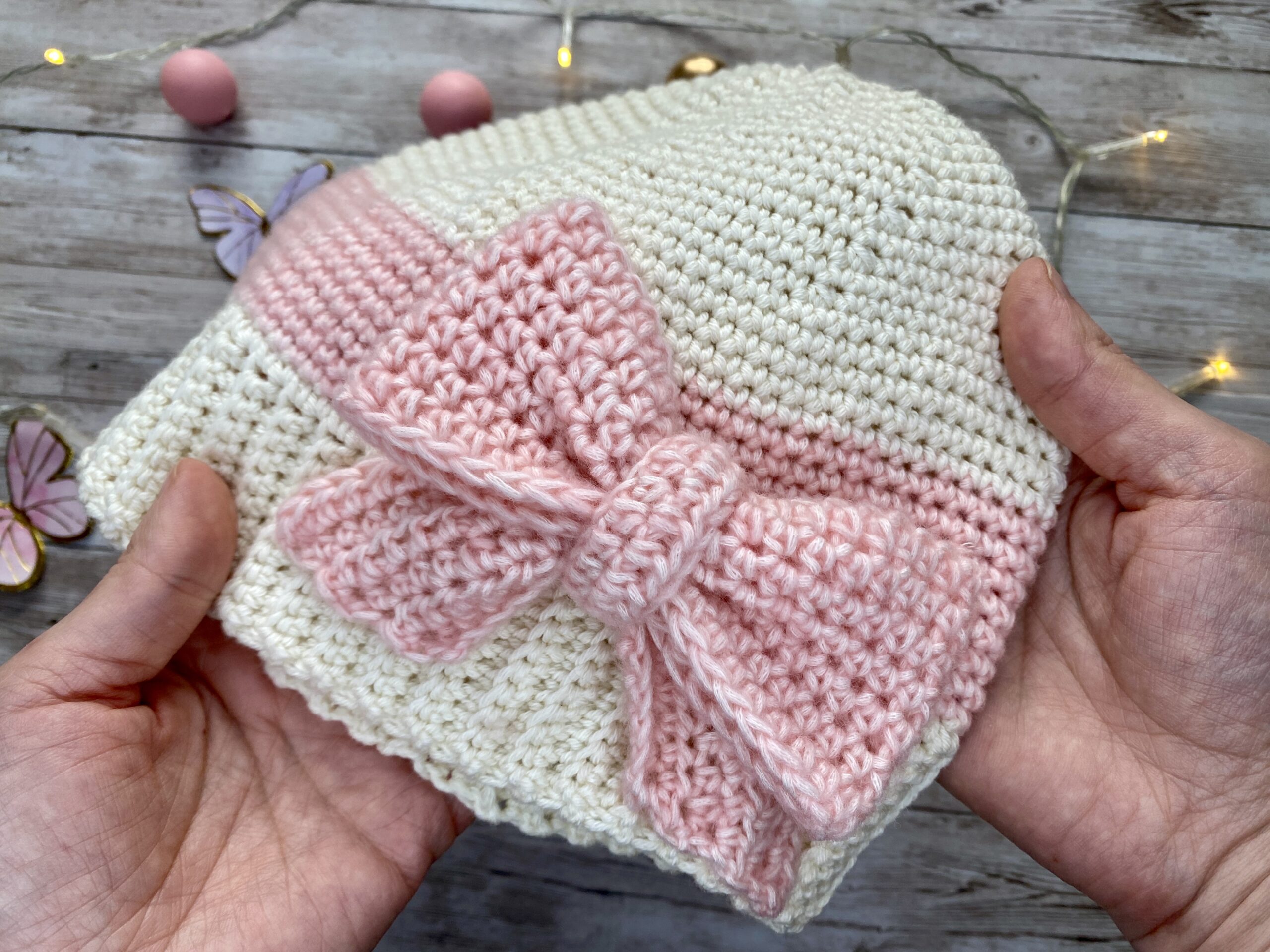 Hand-crocheted cream and blush beanie with a large textured pink bow, shown in close-up to highlight the stitches and bow detail.