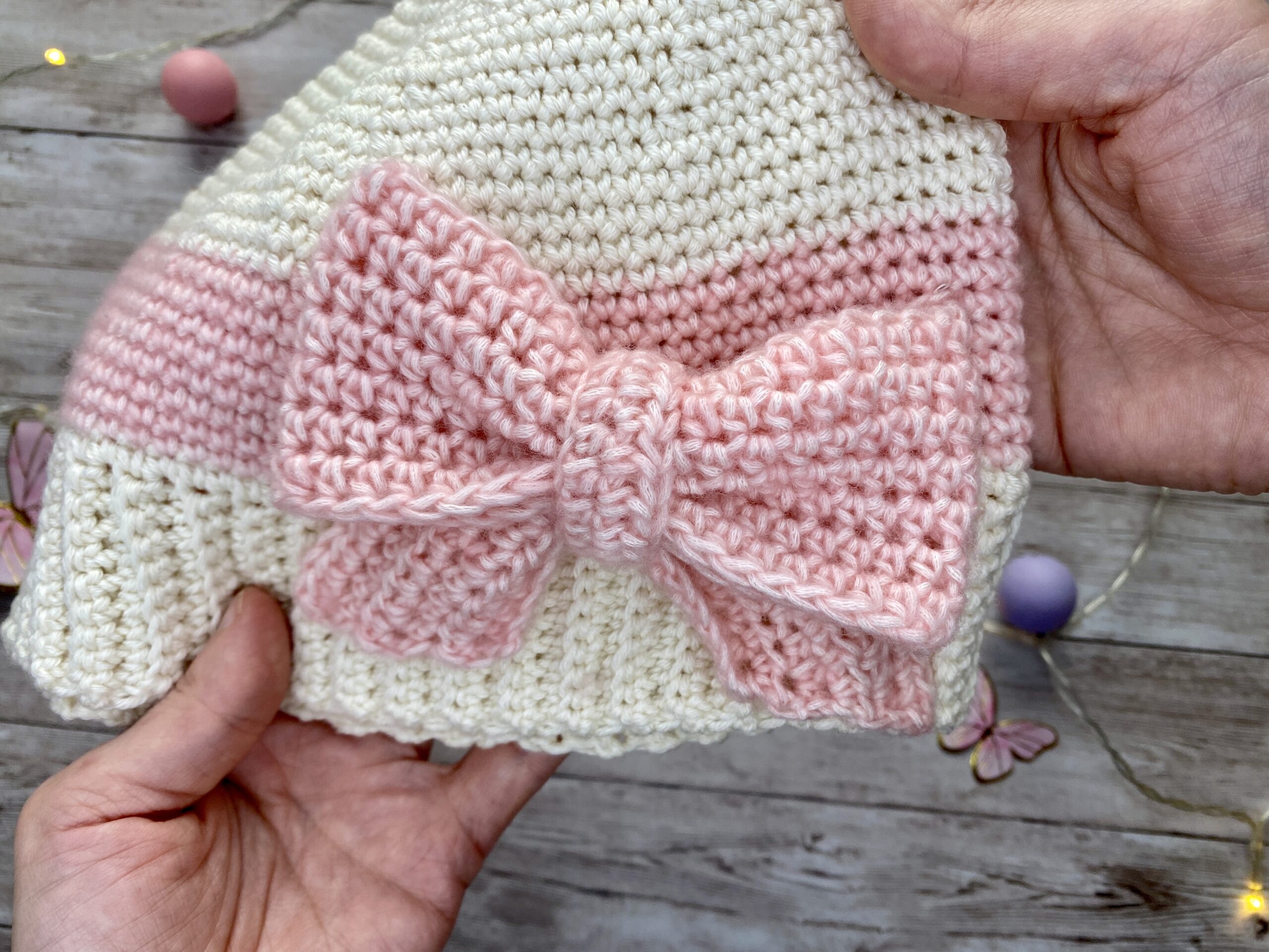 Hand-crocheted cream and blush beanie with a large textured pink bow, shown in close-up to highlight the stitches and bow detail.