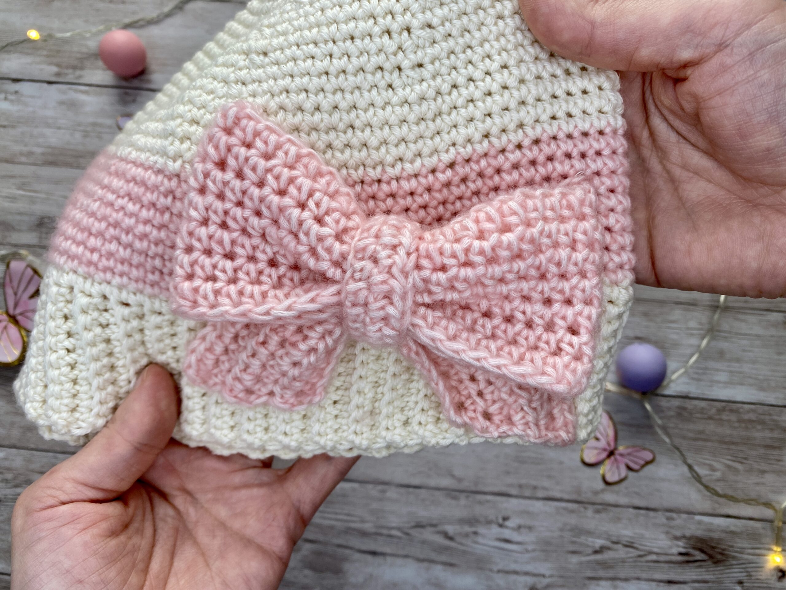 Hand-crocheted cream and blush beanie with a large textured pink bow, shown in close-up to highlight the stitches and bow detail.