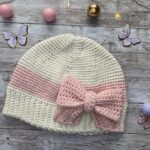 Flat lay of a handmade cream and blush crochet hat with a large pink bow, styled with soft decorative elements in the background.