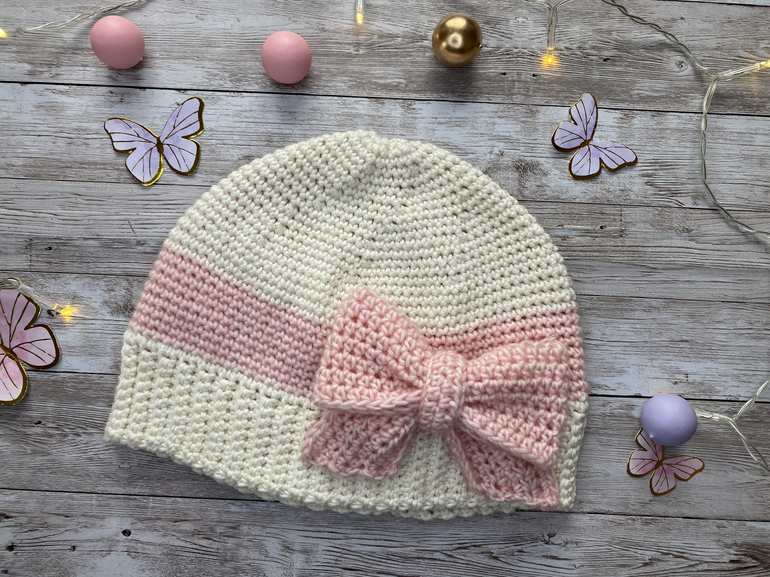 Flat lay of a handmade cream and blush crochet hat with a large pink bow, styled with soft decorative elements in the background.