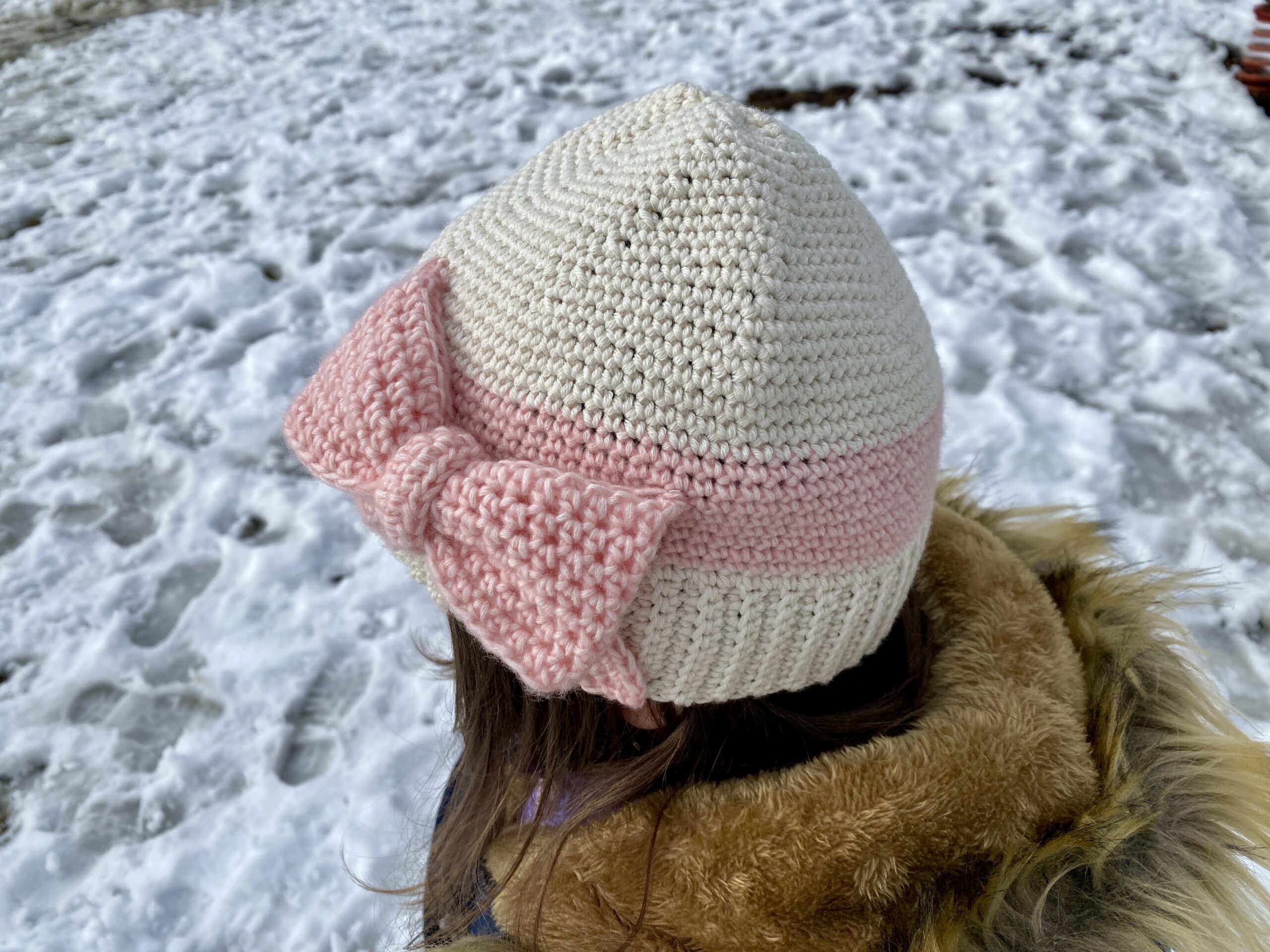 Child wearing a cream and blush crochet beanie with a pink bow on the back, showing how the handmade hat fits and looks when worn
