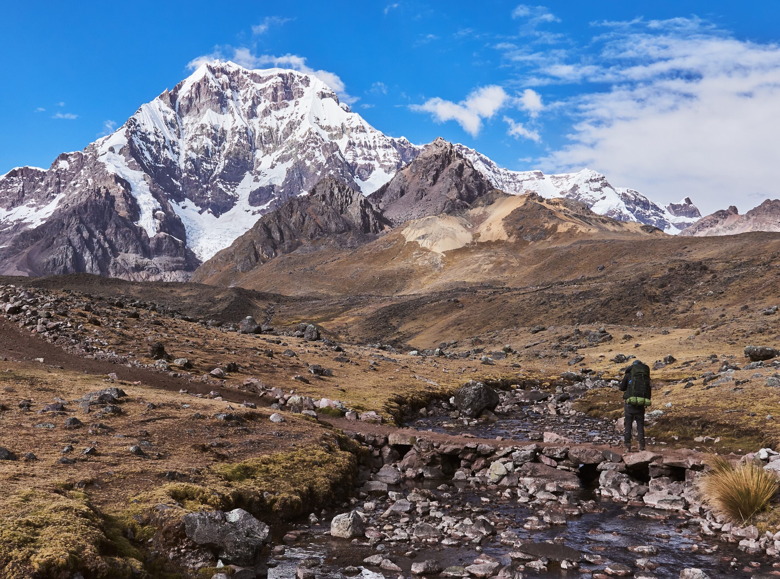 Hiker with backpack crossing a stone bridge in the Andes, with snow-capped peaks in the background