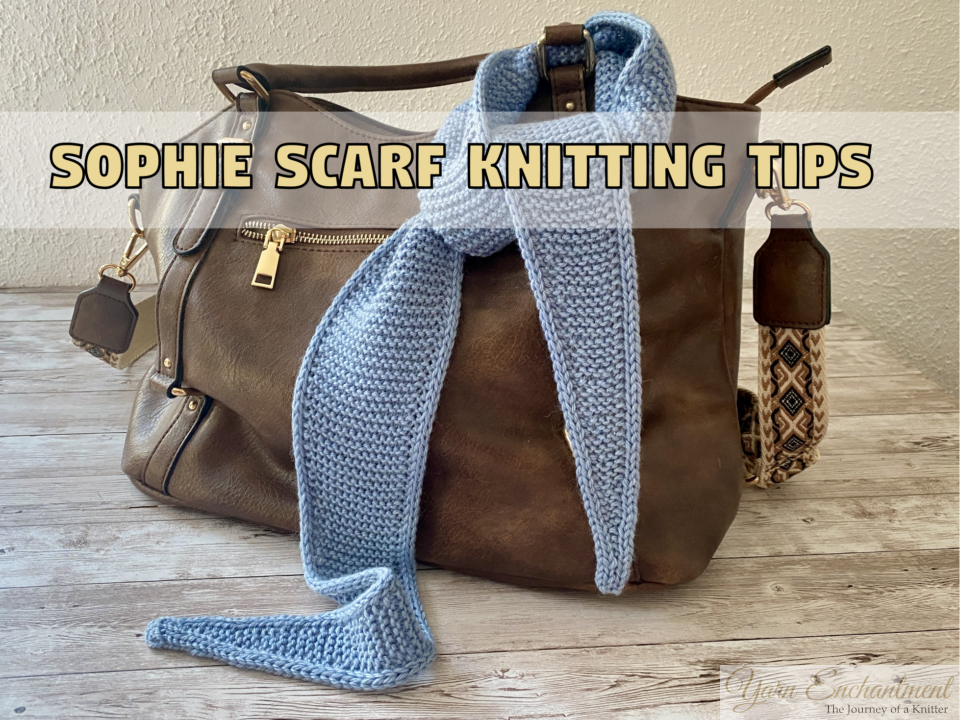 A light blue knitted Sophie scarf draped around the handle of a brown leather handbag on a rustic wooden table. A text banner above the scarf reads "Sophie Scarf Knitting Tips."