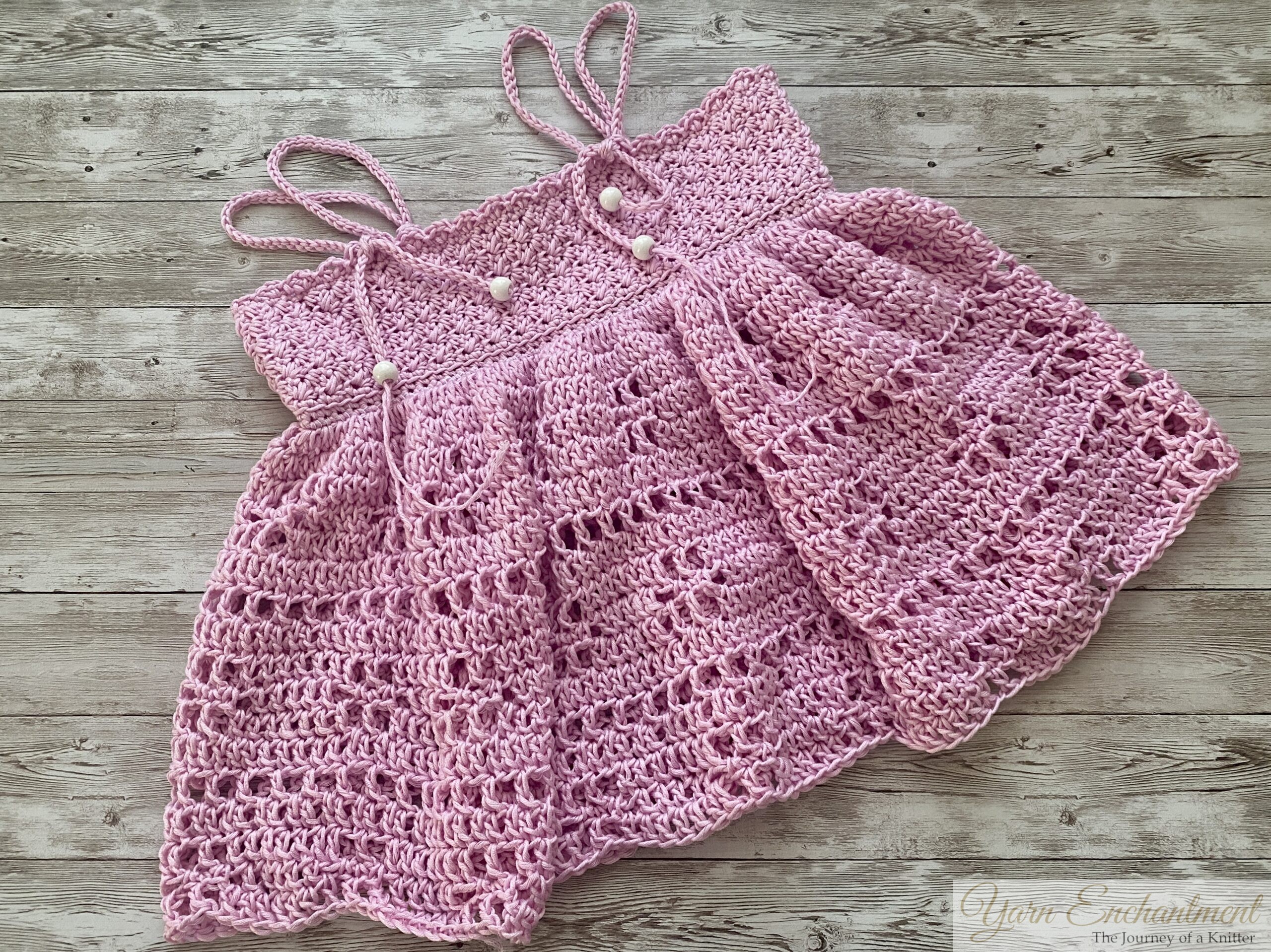 Flat lay of a handmade pink crochet halter top with openwork lace stitching and three decorative buttons, featuring tied shoulder straps and an A-line swing shape, displayed on rustic whitewashed wooden boards.