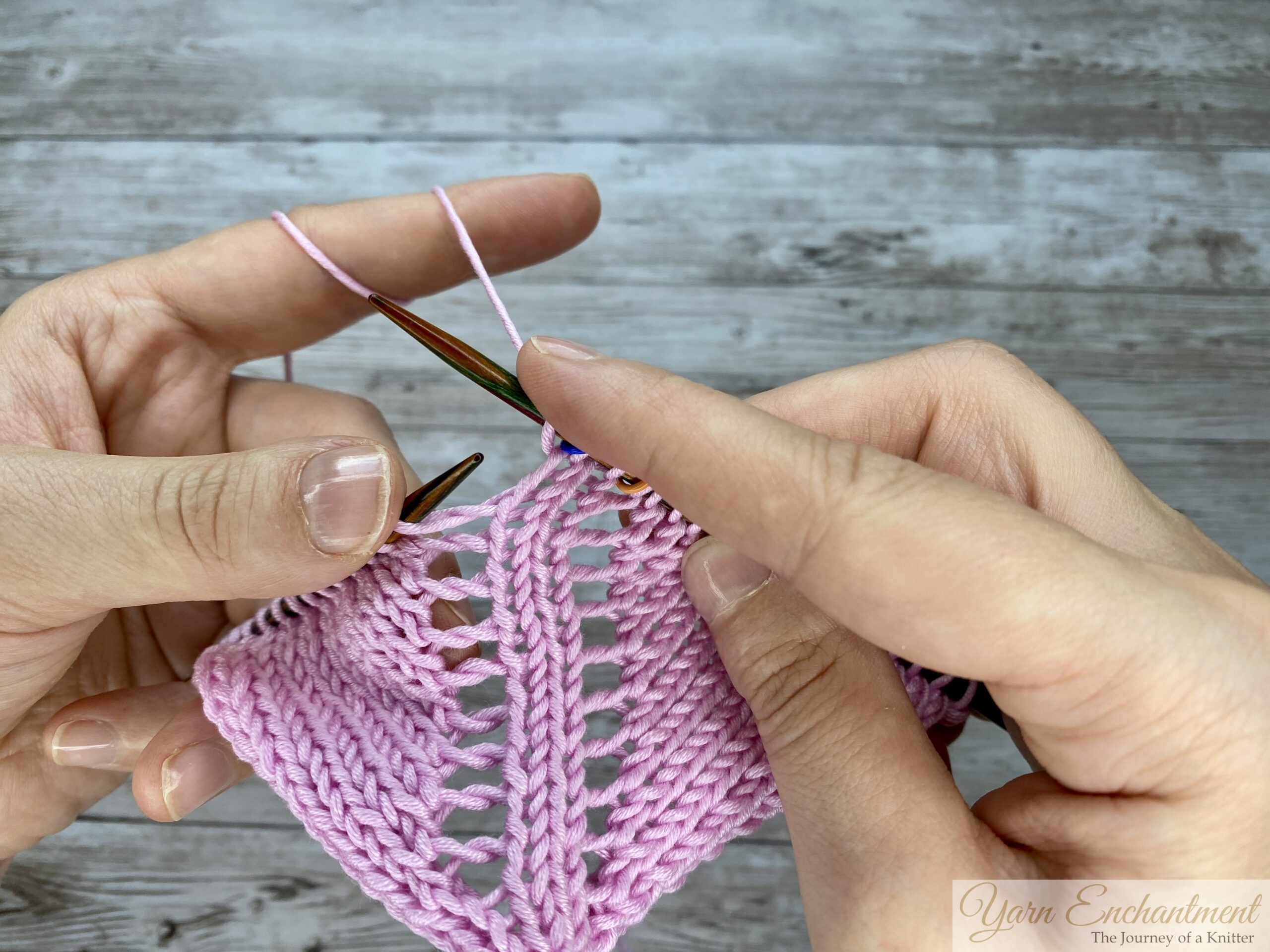 Close-up of hands knitting in the round on circular needles, inserting the right needle into the stitch immediately following a directional yarn-over increase, with a blue stitch marker marking the center point of the pink lace.