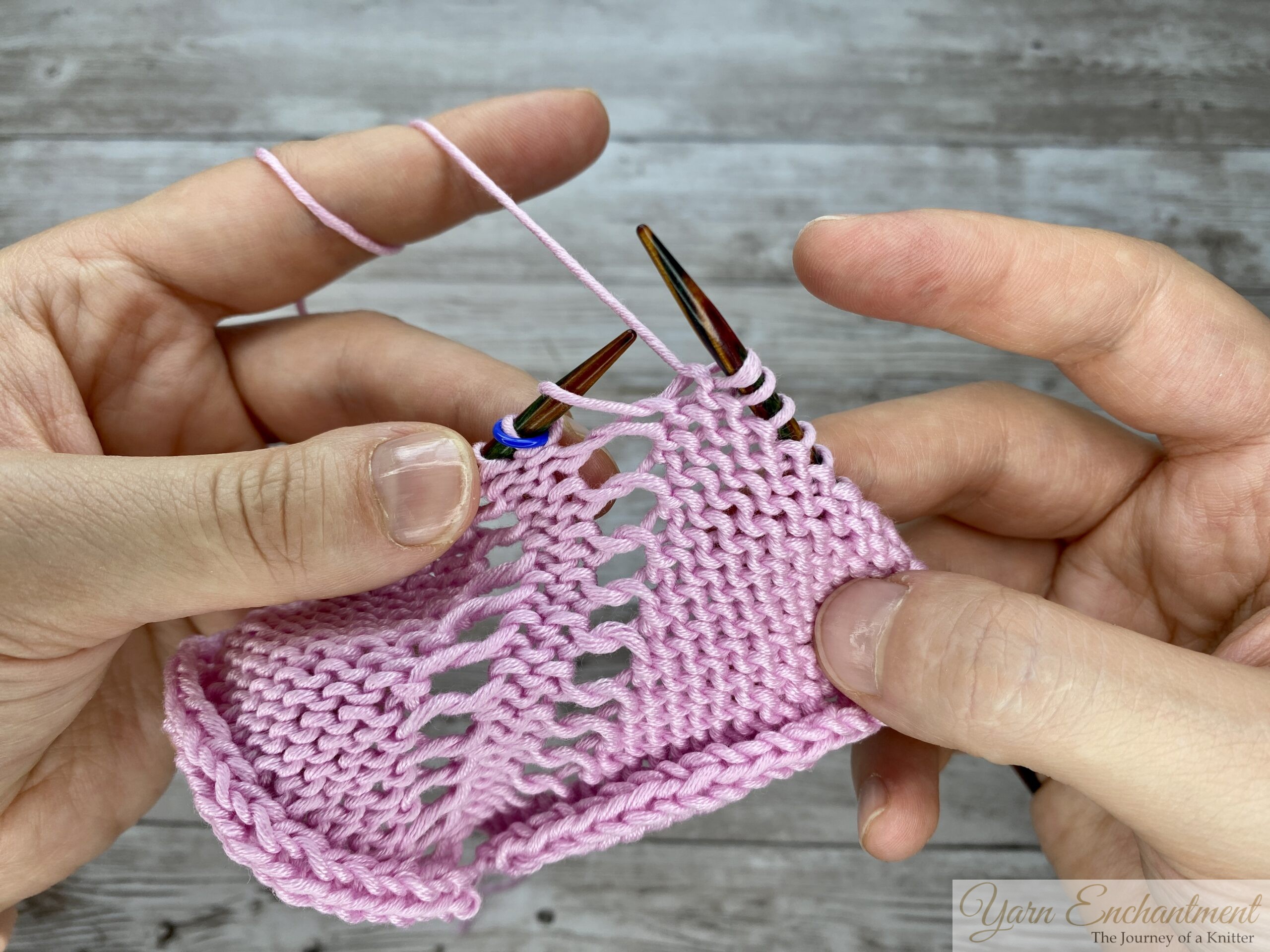 Close-up of hands purling through the front loop on the wrong side to create an open eyelet lace increase, shown in pink yarn on wooden circular knitting needles with stitch markers.