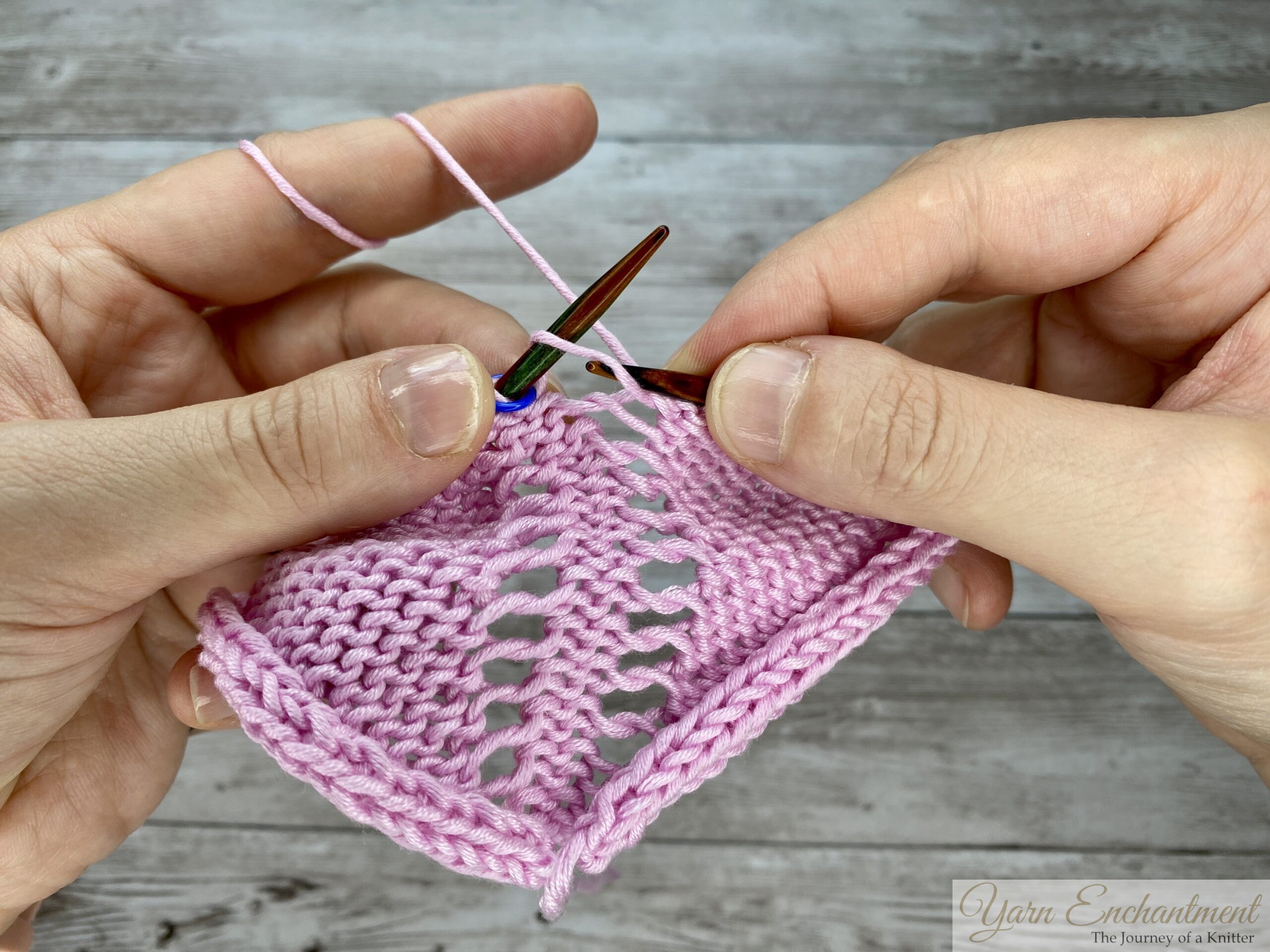 Hands holding pink yarn and circular needles, inserting the needle into the front loop of the stitch to prepare for a purl-increase that creates an open eyelet.