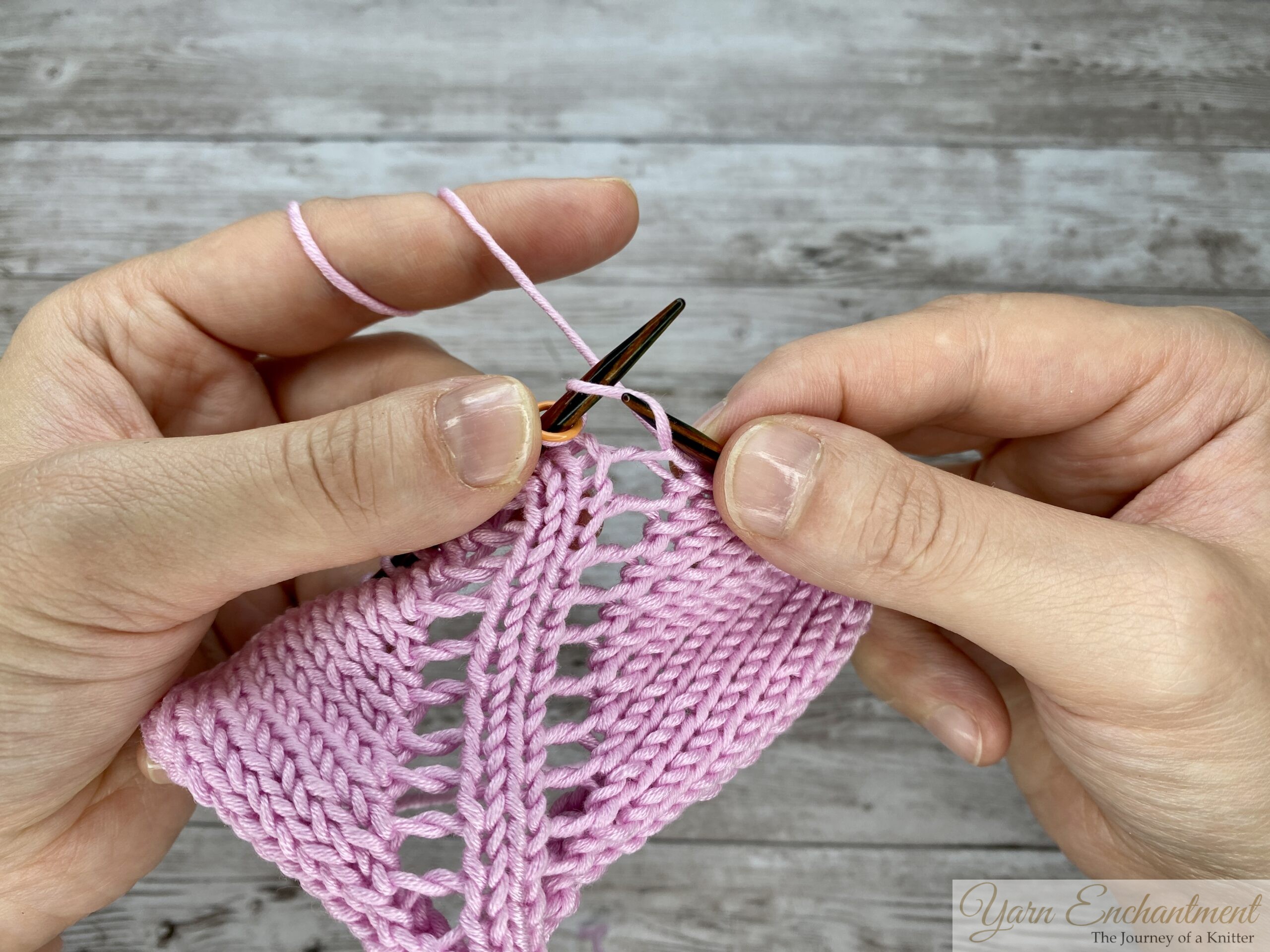 Hands knitting in the round on circular needles, demonstrating a yarn-over increase that creates distinct open eyelet holes in a light pink lace pattern.