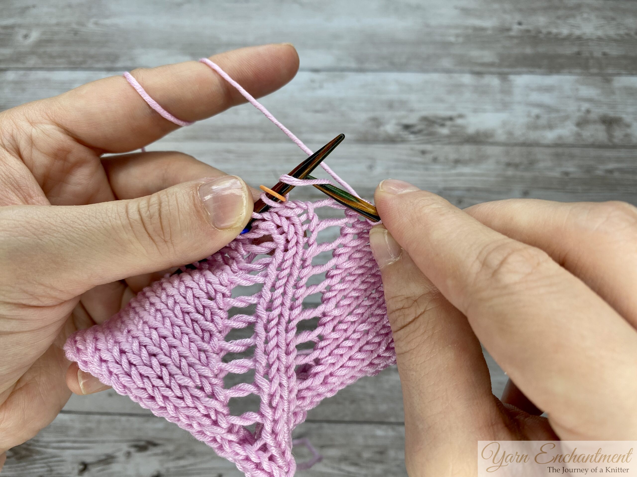 Hands knitting in the round on circular needles, demonstrating a yarn-over increase that creates distinct open eyelet holes in a light pink lace pattern.