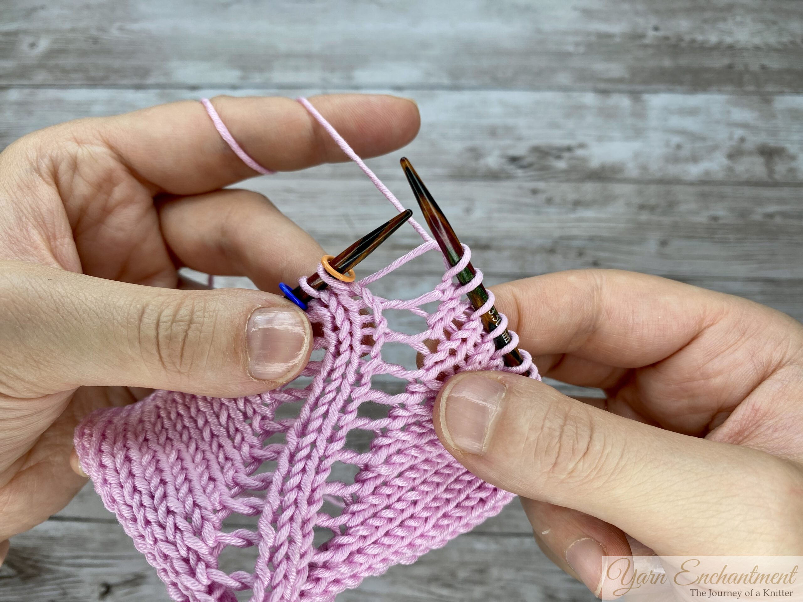 Hands knitting in the round on circular needles, demonstrating a yarn-over increase that creates distinct open eyelet holes in a light pink lace pattern.