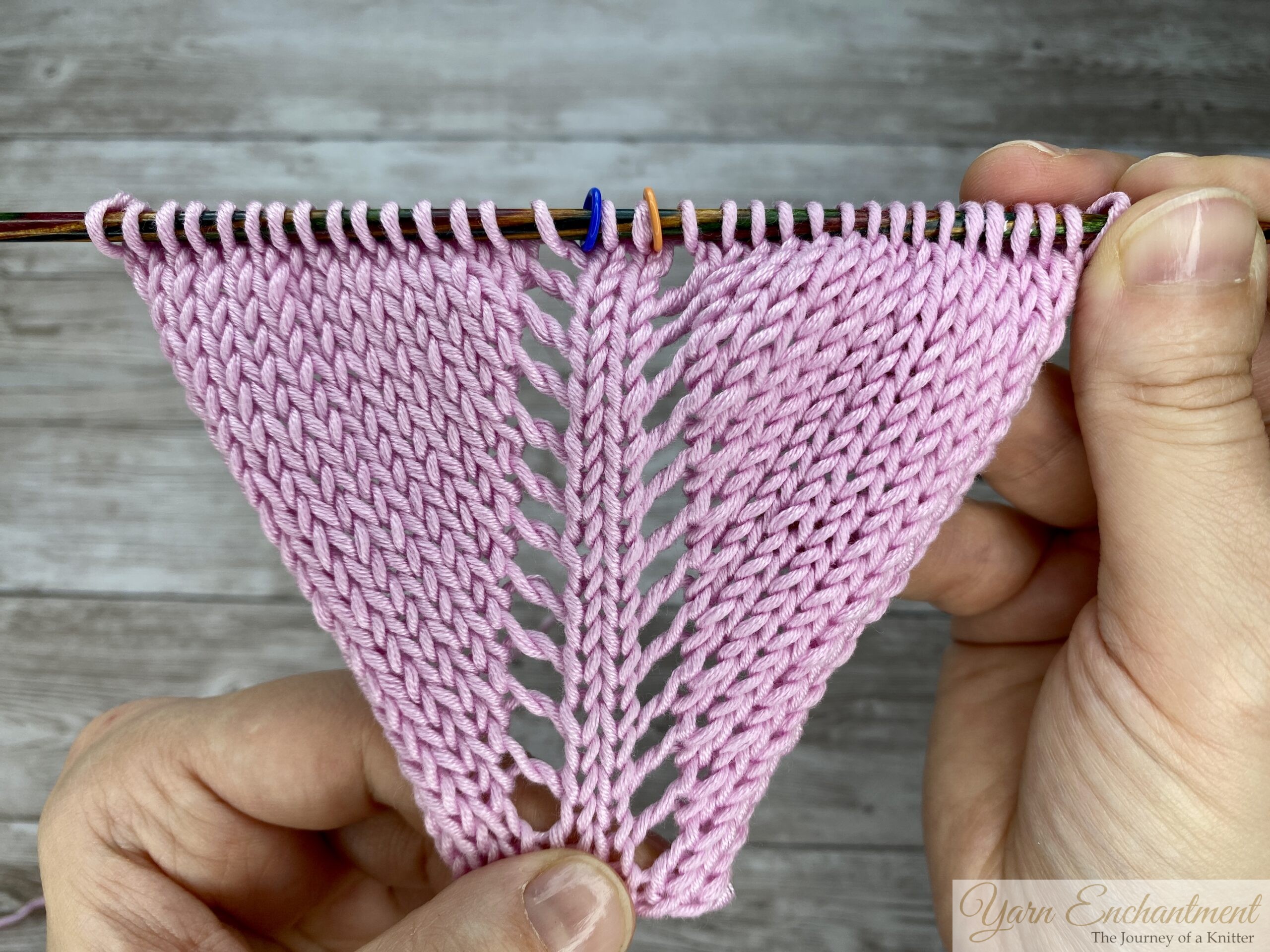 Overhead view of the completed pink garter stitch swatch on a needle, clearly showing the open eyelet lace increases along the centerline between stitch markers.